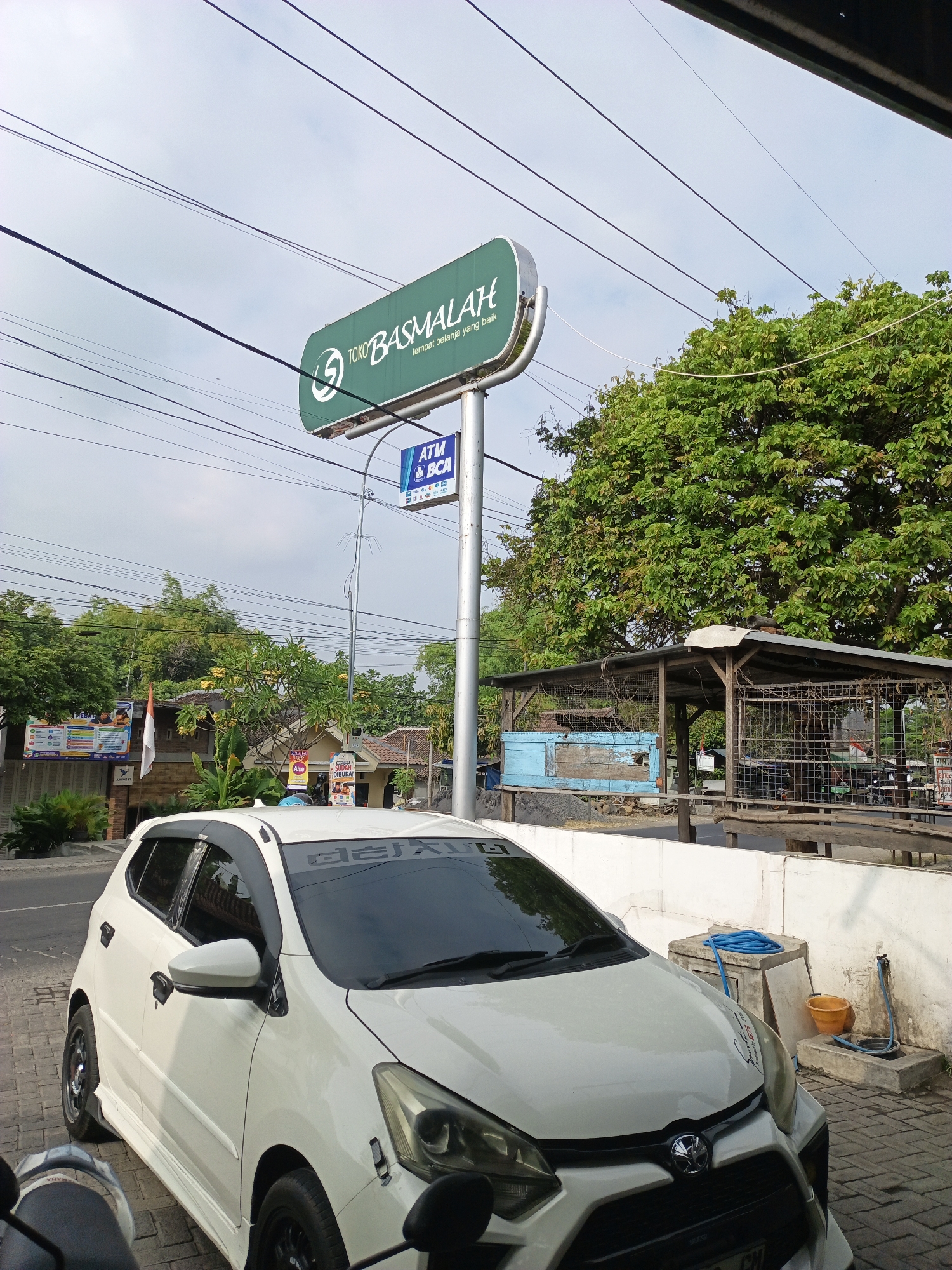 The image shows a white car parked on a paved surface in front of a building with a green sign that reads "TOKO BASMALAH" and "tempat belanja yang baik". Below the green sign, there is a blue sign that reads "ATM BCA". A large green tree is visible behind the signs. The scene appears to be in a commercial area. The location is identified as Baujeng, Indonesia.