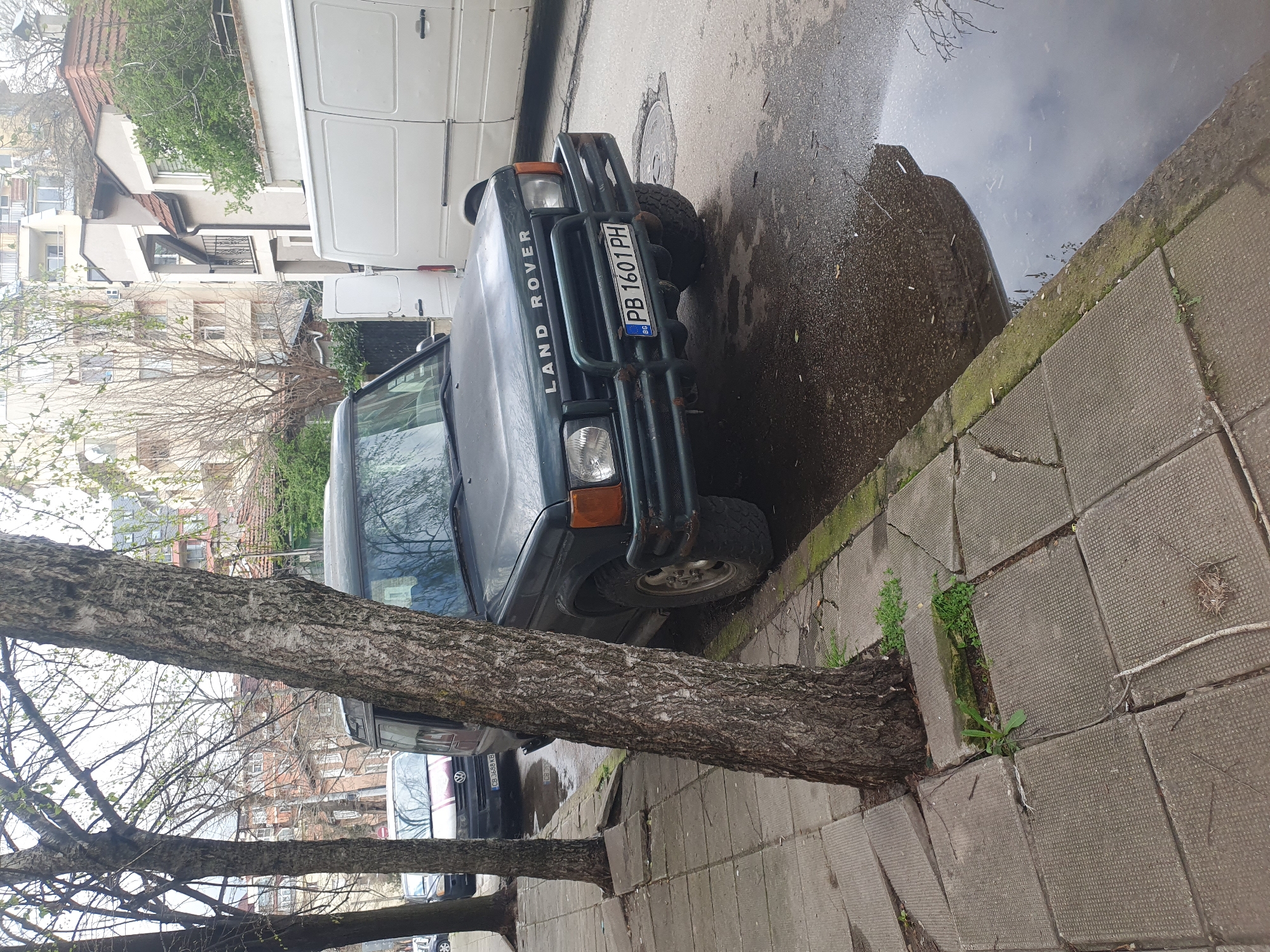 The image shows a green Land Rover Discovery parked on a cracked sidewalk in Plovdiv, Bulgaria. The vehicle has a Bulgarian license plate reading "PB 1601 PH." A tree trunk grows directly out of the sidewalk beside the car. A white van is partially visible behind the Land Rover, and other vehicles are parked further down the street. A large puddle is visible on the pavement. Buildings are visible in the background, including some with balconies. The overall environment suggests an urban, residential area.