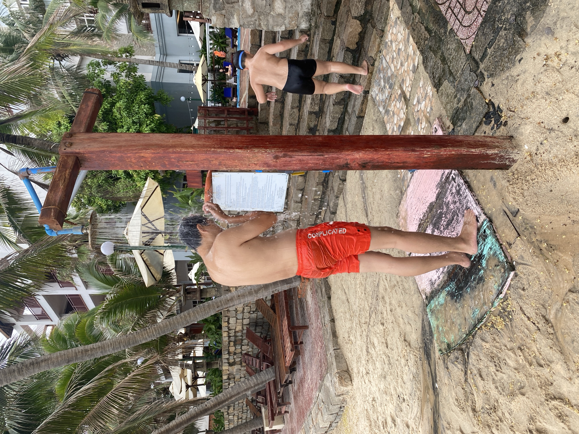 A man in red swim trunks labeled "COMPLICATED" is showering outdoors in Phu Thuy Ward, Vietnam. Another man in black swim trunks is climbing stairs in the background. The scene is a beach area with palm trees and buildings in the distance. The shower is constructed from wood. Water is running.