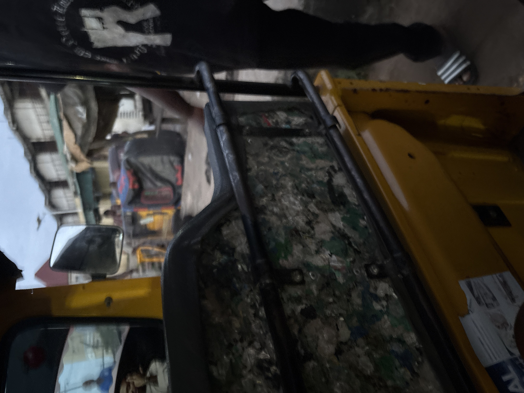 The image captures a close-up view of the interior of a yellow three-wheeled vehicle, likely a commercial motorcycle taxi (okada) or a small delivery vehicle, parked in what appears to be a bustling market area in Onitsha, Nigeria. The foreground is dominated by the vehicle's interior, revealing a black seat with a metallic frame. This frame is filled with a textured material composed of fragmented items, possibly recycled plastic or debris, in shades of green, white, and gray.
To the left, a side mirror reflects a scene outside, showing blurred figures of people and structures. One reflection appears to be a person wearing a blue shirt. Further back, through the vehicle's opening, glimpses of a market environment are visible. This includes stalls with weathered roofing, piled-up bags (some black and red), and indistinct human figures moving about. The overall atmosphere suggests a warm, possibly humid, daytime setting in an urban, commercial district.
On the right side of the image, a person's legs and feet are visible, wearing black trousers and black and white striped sandals. This individual is standing next to the vehicle. The background depicts a row of buildings, some with multiple stories and balconies, suggesting a built-up urban landscape typical of a Nigerian city. The lighting is somewhat muted, possibly indicating overcast conditions or late afternoon.