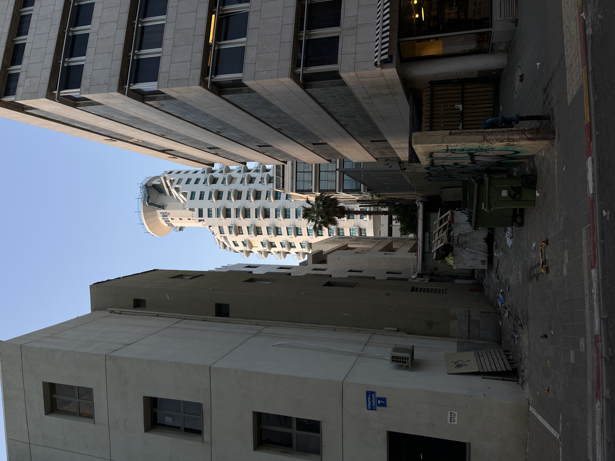 The image captures an alleyway in Tel-Aviv, Israel, framed by tall buildings on either side. The buildings vary in architectural style; the one on the left is characterized by a modern design with protruding rectangular elements and black windows. A curved, white building is also visible further down the alley. The alleyway itself appears narrow and somewhat cluttered, with garbage bins, debris, and a bench along the side. The pavement is paved with gray bricks. A sign with Hebrew characters is visible on the side of one of the buildings. The sky above is clear and blue.