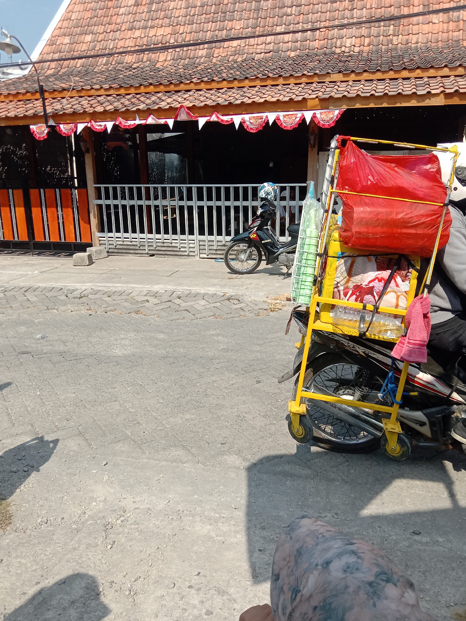 A motorcycle is parked on a paved street in Wirosari, Indonesia, with a large yellow metal rack attached to its rear, carrying a red plastic-wrapped bundle, green plastic cups in a white rack, and two plastic bottles of liquid. A pink towel hangs from the rack. In the background, a building with a tiled roof and a white metal fence is visible. The street is made of concrete pavers. The scene is illuminated by bright sunlight, casting shadows on the ground.