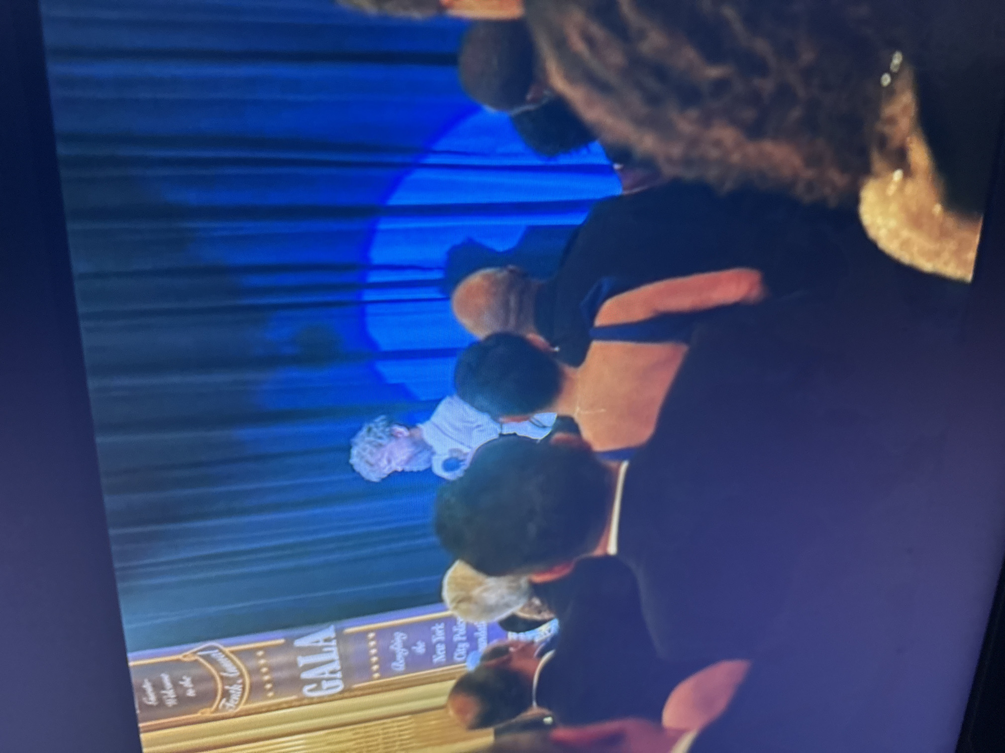 The image shows an event occurring indoors, in an unknown city. A group of people are standing and looking towards the left. The event appears to be a Gala, specifically "Benefiting the New York City Police Foundation". A blue backdrop is present, with lighting illuminating the scene.