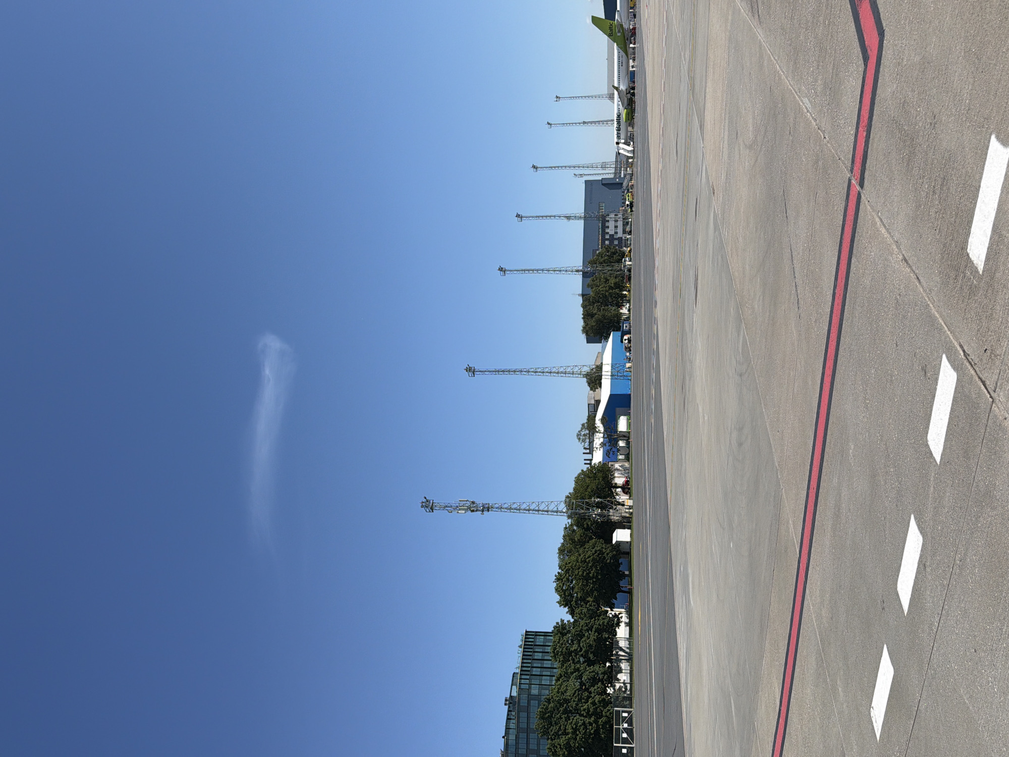 The image shows an outdoor scene at an airport on a clear day. In the foreground, there is a concrete tarmac with white and red markings. In the middle ground, there are several tall communication towers, trees, buildings, and an airplane with the livery of "airBaltic" parked at a gate. The background consists of a clear blue sky with a faint cloud formation. The scene suggests an operational airport environment, likely in Tallinn, Estonia, given the presence of the airBaltic airline which is the flag carrier of Latvia and is headquartered in Riga, but also serves Tallinn.
