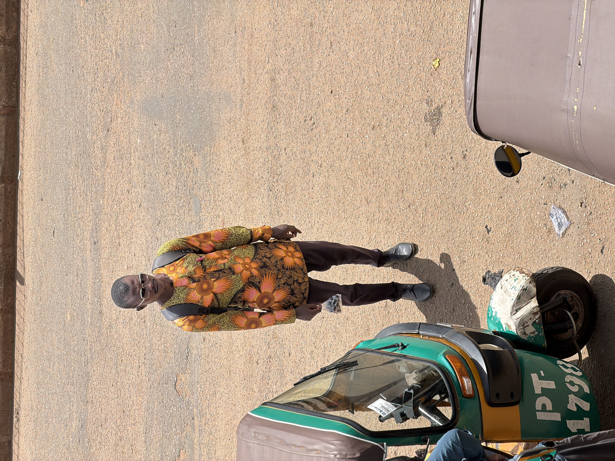In Bukuru, Nigeria, a man wearing a vibrantly patterned orange and yellow shirt, dark trousers, and sunglasses stands on a dusty, asphalt road. He carries a black backpack and holds a small plastic bag filled with dark objects in his right hand. To his left, a green and yellow auto-rickshaw, identified by the markings "PT-1798" on its side, is parked. Another vehicle, partially visible and brown in color, is positioned in the lower right corner of the frame. The environment is characterized by a dry, sandy road surface and the bright light of a sunny day, casting distinct shadows.