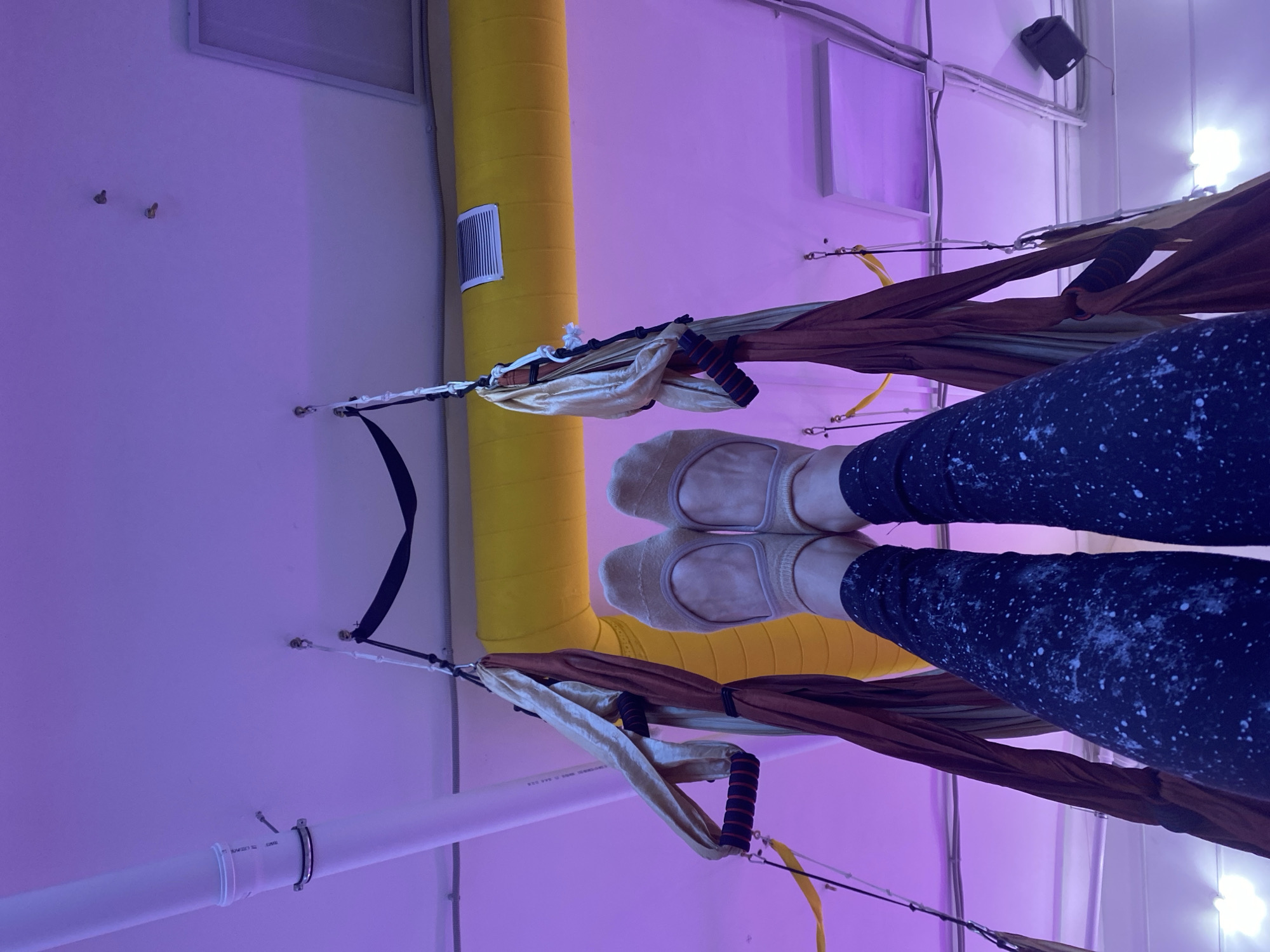 The image presents a low-angle perspective of a person suspended in an aerial yoga hammock, presumably indoors. The individual is wearing navy leggings with a white speckled pattern and light beige socks. Their legs are positioned horizontally within the hammock, which is composed of brown and beige fabric. The hammock's handles are visible, featuring black and red grips.
The background consists of a light purple wall and ceiling. A thick, yellow cylindrical object, likely a padded support, is visible behind the hammock. There are white pipes and various fixtures attached to the ceiling and wall, along with a black speaker.
Given the information provided, the image does not contain enough information to accurately place the image in Chaikovsky, Russia.