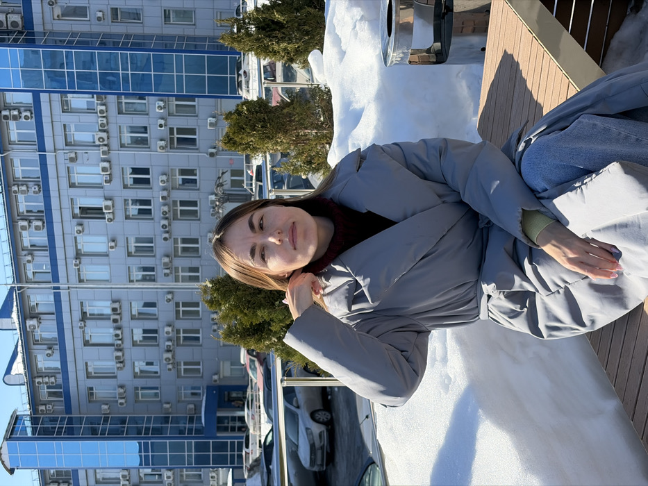 The image displays an outdoor scene in Izhevsk, Russia, on a clear, sunny day with visible snow cover. The location is identified as the area adjacent to the building at Kirova Street 111, Izhevsk.
An adult female individual is seated in the foreground on a light brown slatted outdoor bench with a metallic frame. She has light brown hair and a neutral facial expression, looking slightly to her left. Her right hand is raised, with fingers positioned near her right ear, appearing to adjust hair or an ear accessory; her fingernails are painted light pink. She is dressed in a light grey padded, belted coat, a dark red turtleneck, and light blue jeans, with a light green cuff visible on her left wrist.
To her immediate left, a significant snowdrift is present on the ground. Behind her, a series of small, conical evergreen shrubs are visible, some partially covered in snow. Further back, a parking area contains multiple parked vehicles, predominantly light-colored cars. The background is dominated by a multi-story modern building, identified as part of the complex at Kirova Street 111. This building features grey facades with numerous rectangular windows and a prominent vertical section constructed from blue glass panels. Multiple air conditioning units are affixed to the building's exterior below various windows. A cylindrical metallic object, possibly a trash receptacle or outdoor heater, is partially visible to the right of the bench. The scene is illuminated by strong sunlight, casting distinct shadows.