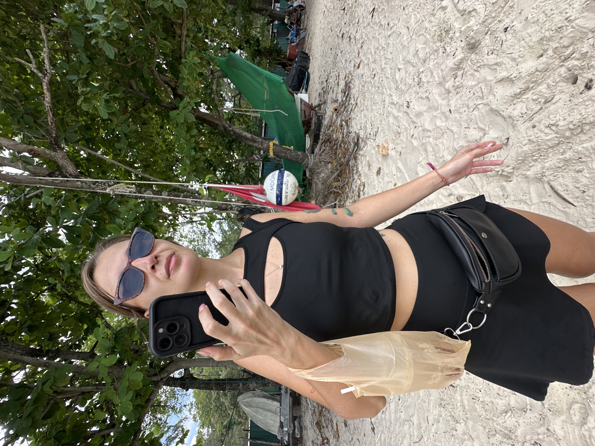 A woman stands on a sandy beach in Karon, Thailand, holding a smartphone in her right hand and a plastic bag in her left. She is wearing sunglasses, a black crop top with a cutout at the chest, and a black skirt. A black fanny pack is worn around her waist. The beach is lined with trees, and in the background, there are boats, a green tarp, and beach chairs.