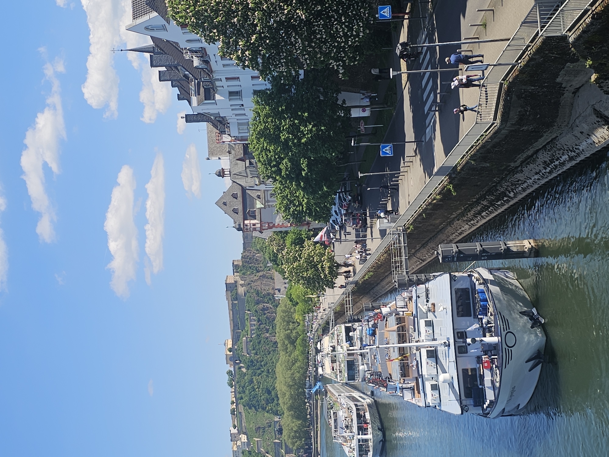 The image shows a view from above of the Rhine River in Koblenz, Germany. Several tourist boats are docked along the riverbank. The Ehrenbreitstein Fortress is visible on a hill overlooking the river. A pedestrian walkway runs parallel to the river, where people are walking. Buildings and trees line the opposite side of the walkway. The sky is blue with scattered white clouds.
