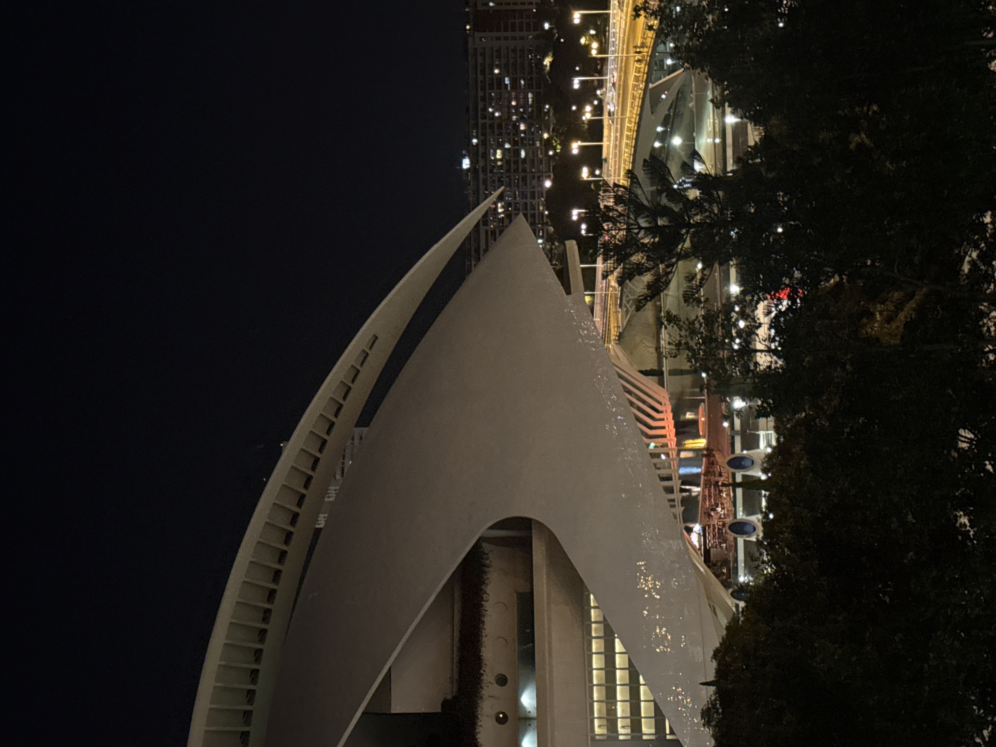 The image captures a nighttime view of the Hemisfèric, a building in the City of Arts and Sciences in Valencia, Spain. The iconic structure, resembling a giant eye, is illuminated, with its white, curved architecture contrasting against the dark sky. Below the Hemisfèric, a bridge with streetlights is visible, with cars crossing. In the background, a city skyline with numerous illuminated windows suggests a residential area. The foreground is dominated by dark, silhouetted trees, indicating a vantage point from a park or elevated position overlooking the scene. The overall impression is one of urban nocturnal beauty, highlighting modern architecture against a backdrop of city lights.