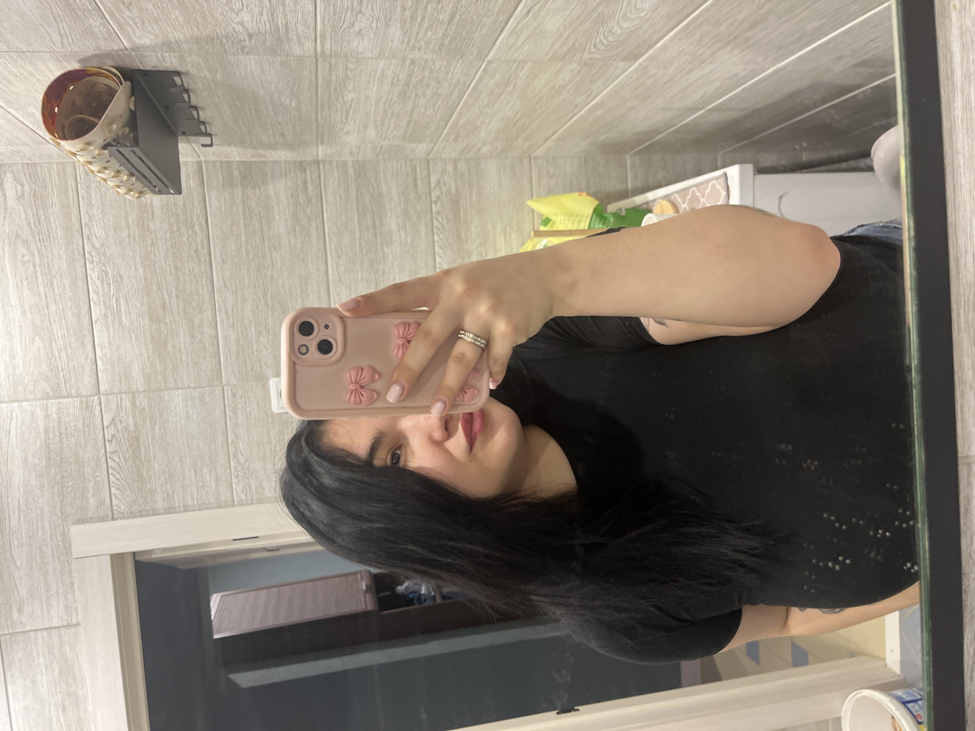 A female individual is depicted in a mirror selfie within an indoor setting, likely a bathroom or utility room. She has long, dark hair and is wearing a black t-shirt and blue jeans, with a tattoo partially visible on her left upper arm. Her left hand holds a pink smartphone featuring a decorative case with bows and a triple-lens camera array; a multi-band ring is present on her left ring finger, and her fingernails are painted pink.
The environment features walls clad in light grey, wood-grain patterned rectangular tiles. To the left, a light-colored wooden door frame opens to a darker room containing brown furniture. On the tiled wall to the right, a black metal shelf with a railing and four hooks is mounted; it holds a cream-colored, ornate container with rolled items. Below the shelf, a white cabinet or vanity with a patterned mat on top is partially visible, with a yellow object resting behind the individual's right shoulder. A white plastic bucket with colorful labels is on the floor or a low surface to the lower left.
The specific location within the city of Chaikovsky, Russia, is not discernible from the provided image.