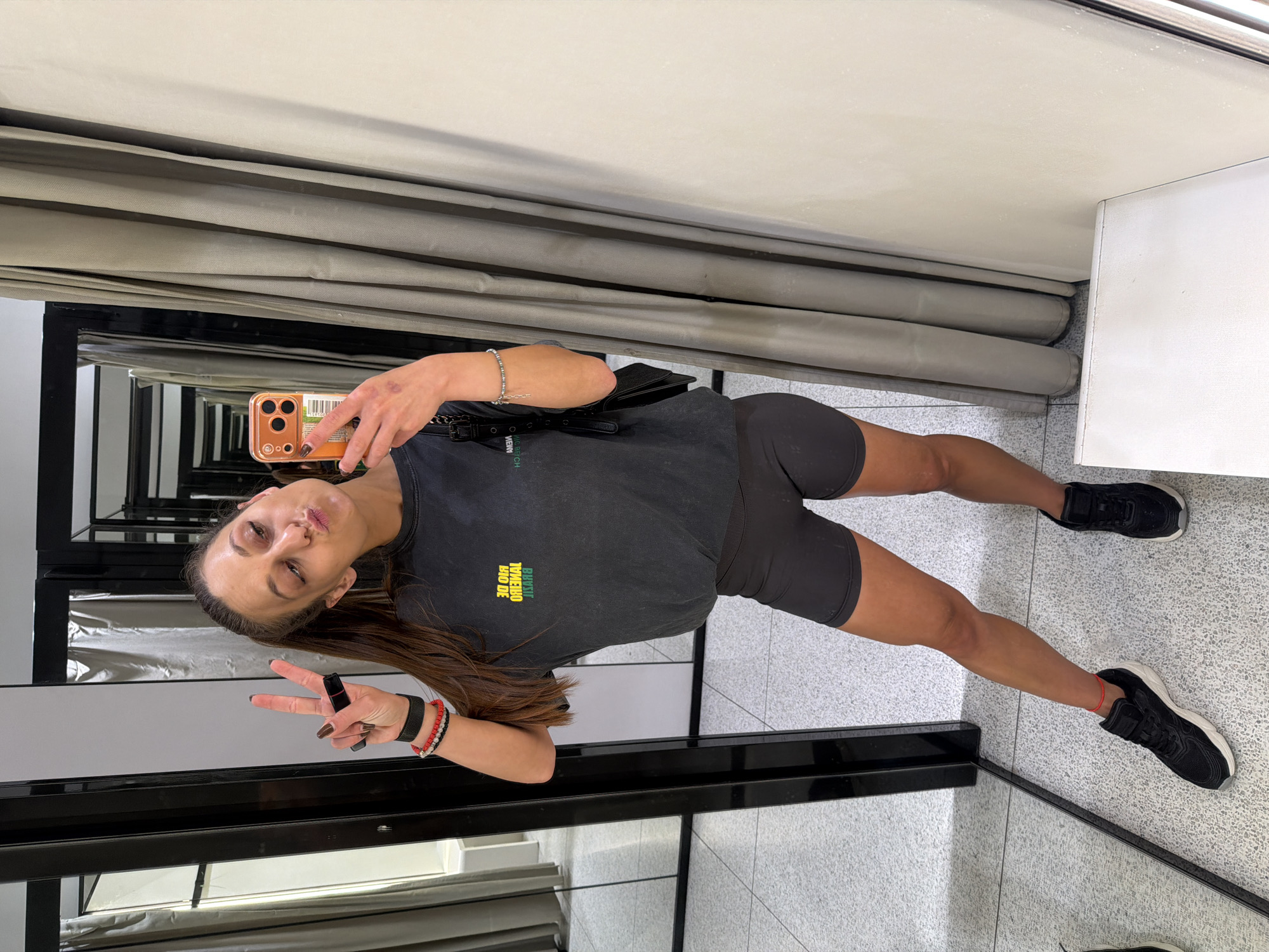 A woman is taking a mirror selfie in what appears to be a fitting room. She has her hair in a high ponytail and is wearing a dark gray t-shirt with yellow and green lettering on the front, and dark gray athletic shorts. She is also wearing black athletic shoes with white soles, and multiple bracelets on her left wrist. She is holding her phone in her right hand, which is an orange case. Her left hand is held up with two fingers extended in a peace sign. The fitting room has tiled flooring, mirrors, and gray curtains. There are no specific indicators in the image that place this scene in Plovdiv, Bulgaria.