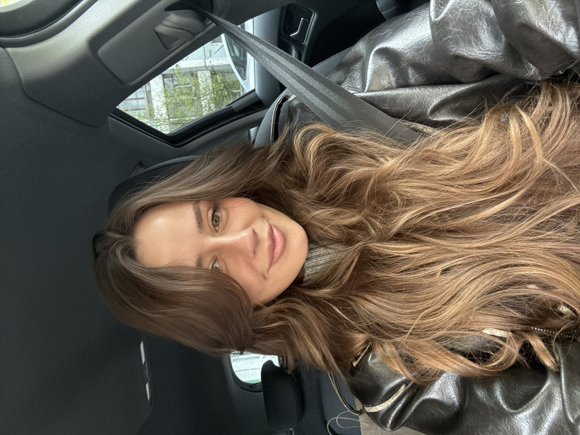 A young woman with long brown hair is sitting in the passenger seat of a car. She is wearing a seatbelt and a grey turtleneck sweater under a black jacket. The car has a sunroof, and trees and a building can be seen through it. The interior of the car is black. The woman is smiling at the camera. There is no location information available.