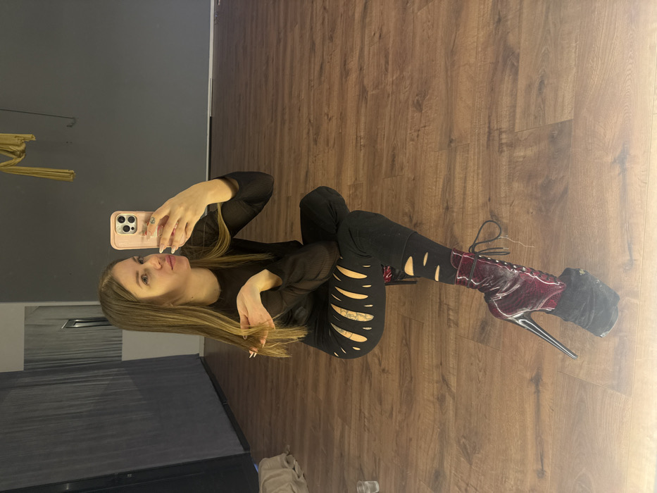 An indoor studio space in Batumi, Georgia, is depicted. A female subject with long, straight, light brown hair is seated on a light brown laminate wood floor, taking a mirror selfie. She wears a black, long-sleeved ribbed top and black leggings with numerous horizontal slits revealing skin on the thighs and knees. Her visible right foot is clad in a dark red, patent leather, lace-up ankle boot featuring an extremely high heel and a prominent black velvet-like protective covering over its platform. She holds a light pink-cased smartphone in her raised right hand, aiming it towards her reflection. Her left hand is partially raised, fingers slightly curled, near her hair. The background comprises a plain dark grey wall. A large mirror on the left reflects a dark grey curtain or partition and a segment of a white wall. In the upper right portion of the background, a piece of yellow fabric, possibly aerial silks, is suspended from the ceiling. A small, clear drinking glass or cup rests on the floor in the lower-left foreground.
