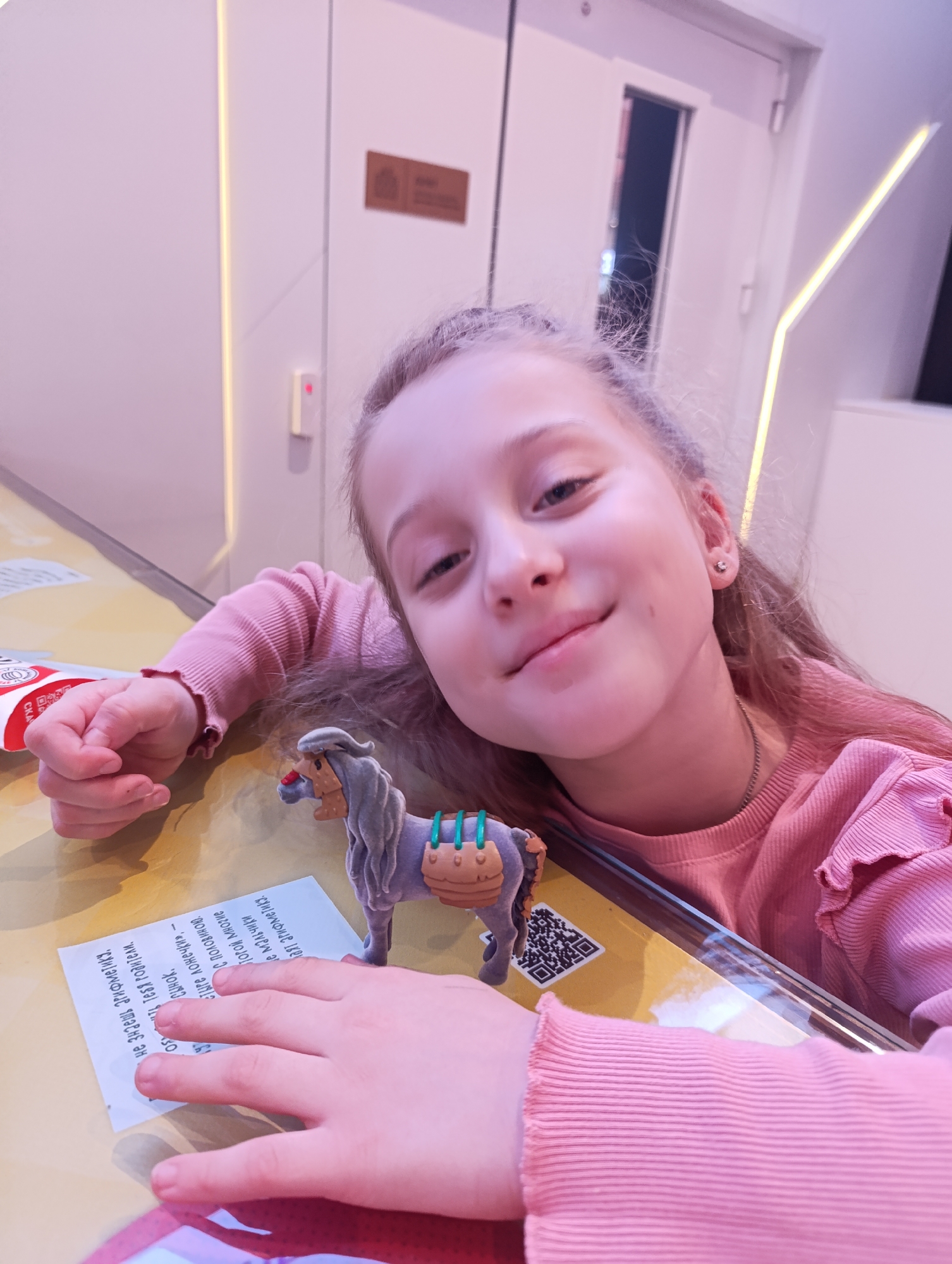 A young girl with light brown hair and a pink long-sleeved shirt is posing for a picture at a table. She is smiling slightly, and her head is resting on her hand. On the table, in front of her, is a small toy horse, a sheet of paper with text in Cyrillic, and a red and white container. The QR code is visible on the table. The location is likely Moscow, Russia, based on the Cyrillic text.
