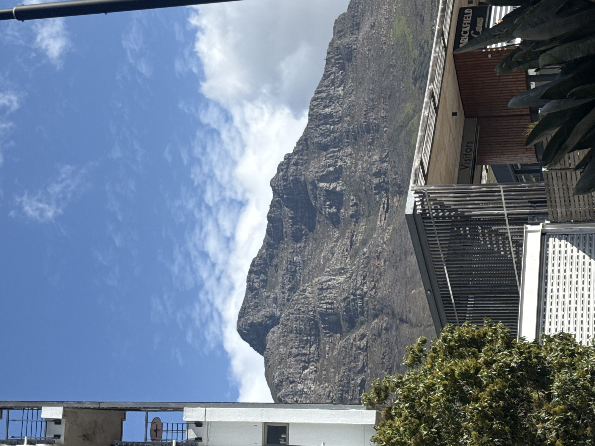The image shows a view of Lion's Head mountain in Cape Town, South Africa, taken from an elevated position. The mountain dominates the right side of the frame, rising sharply against a sky with scattered clouds. The bottom of the frame includes rooftops, and the middle ground shows vegetation and sections of the building, identified as "Brickfield Canvas Visitors". The lighting indicates a sunny day, and a black object is present on the top left. The scene suggests an urban environment close to natural landmarks.