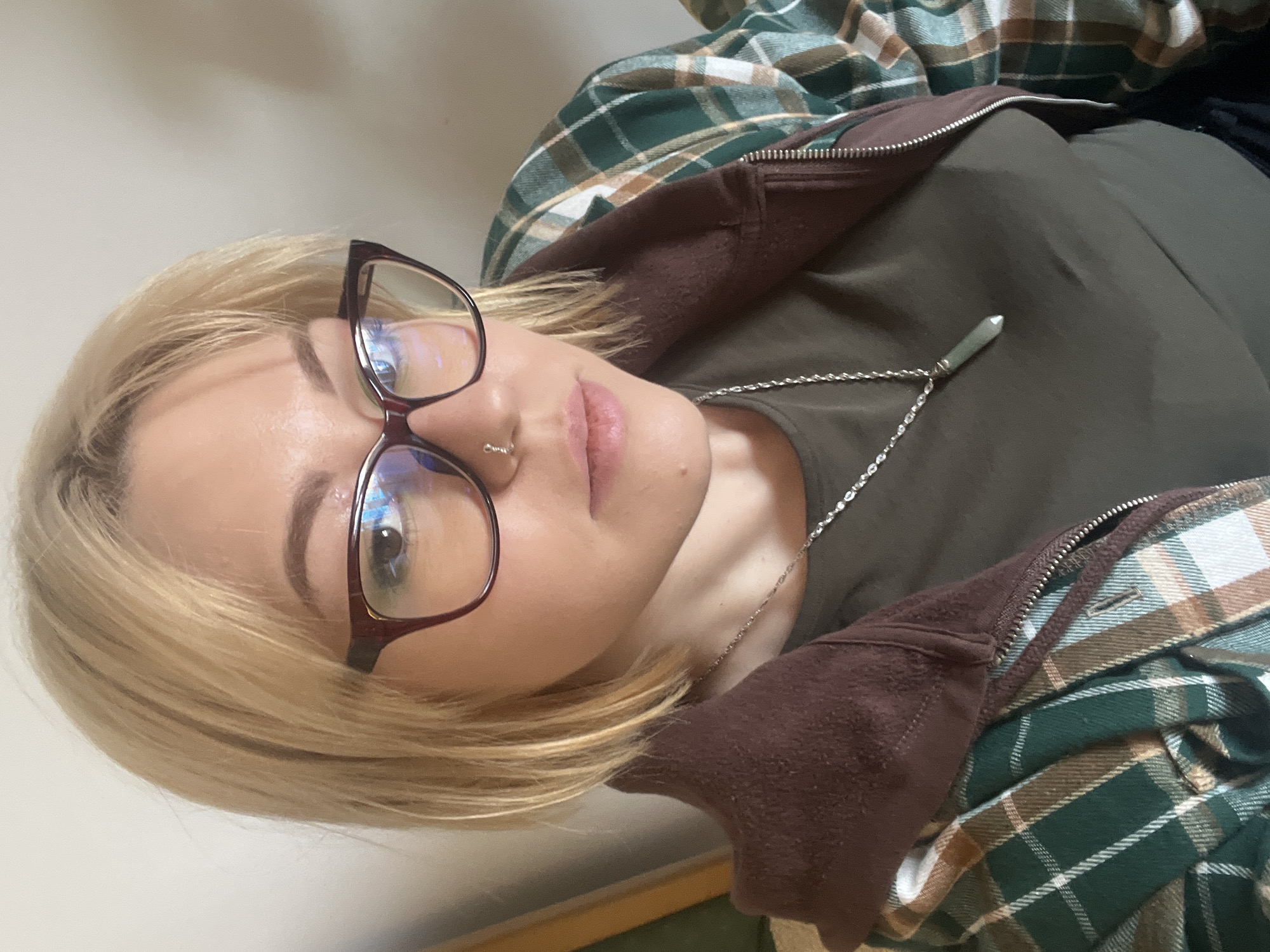 An eye-level portrait shows a person with blonde hair and fair skin, wearing cat-eye style glasses with dark frames and a small silver hoop nose ring on their left nostril. The subject has a neutral expression, looking directly towards the camera. They are dressed in layers, including a dark olive green crew-neck t-shirt, a partially unzipped brown hooded jacket or sweatshirt, and an unbuttoned green, brown, and white plaid shirt worn over the jacket. A silver chain necklace with a light green, pointed, crystal-like pendant rests against the green t-shirt. The background consists of a plain, light-colored wall, with a glimpse of a darker surface at the bottom right. The image is oriented sideways, implying the subject is reclining or the photo was taken from an angle. There is no visible text. The location context for this image is Budva, Montenegro.