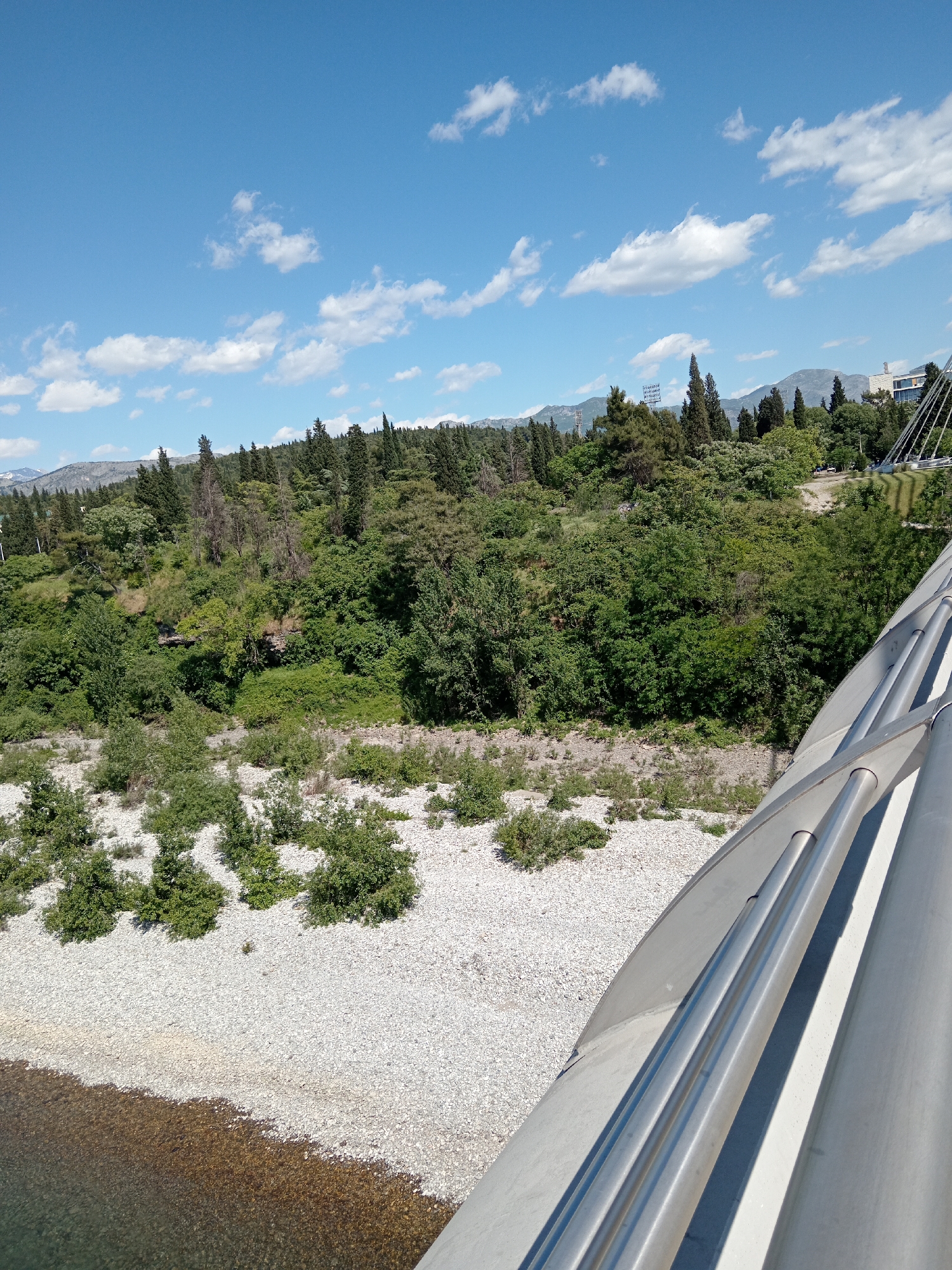 A high-angle, wide shot captures a sunny day overlooking a riverbed in Podgorica, Montenegro. The riverbed is filled with grey gravel and sparse, green vegetation leading to a dense forest on the far bank. A body of water, likely the Moraca River, is visible at the bottom left. In the background, hills and mountains are visible under a bright blue sky scattered with white clouds. The shot is taken from a bridge; part of the bridge's metallic structure dominates the right side of the frame.