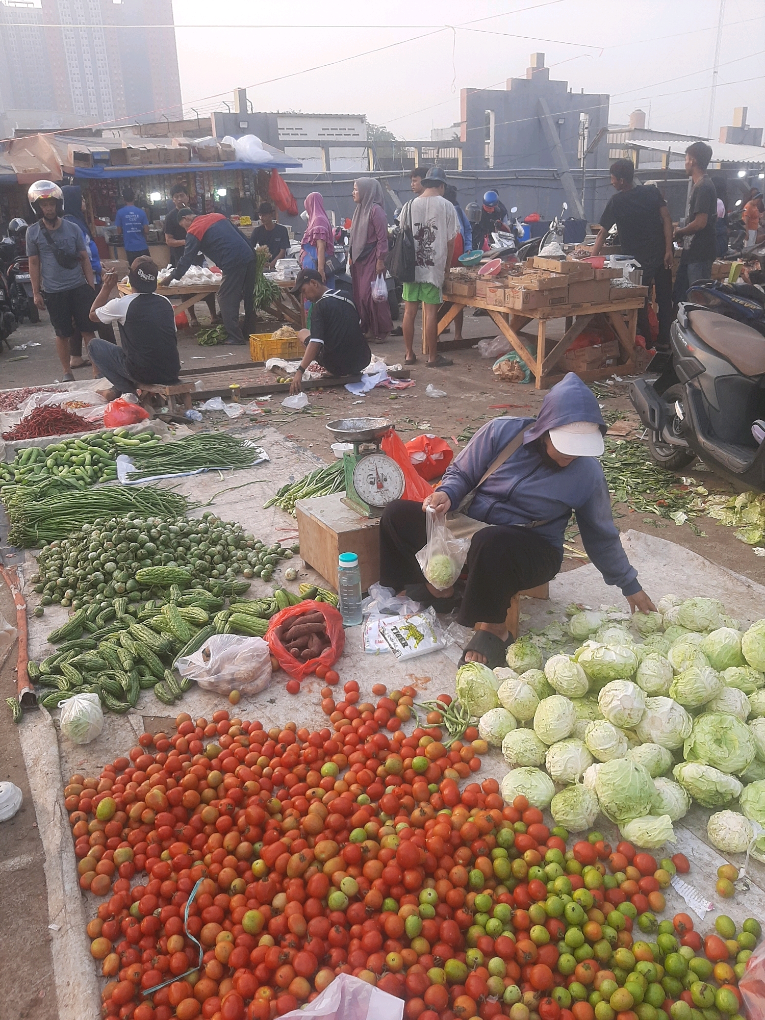 This image depicts an outdoor market scene in Bekasi, Indonesia. The foreground is dominated by a large display of fresh produce, including piles of tomatoes, cabbages, long beans, cucumbers, and what appear to be small eggplants. A person wearing a blue hoodie and a white cap is seated on a stool, holding a plastic bag with cabbage and reaching towards other cabbages on the ground. To the left, a scale is visible, along with red chili peppers and other assorted vegetables.
In the background, several individuals are seen engaged in market activities. Some are standing and interacting, possibly as vendors and customers, while others are seated. There are also market stalls and structures, suggesting a bustling commercial area. Motorcycles are parked in the vicinity, a common sight in Indonesian markets. The overall environment appears to be a busy, open-air marketplace with goods spread out on tarpaulins and tables. The sky is overcast, indicating potentially hazy weather.