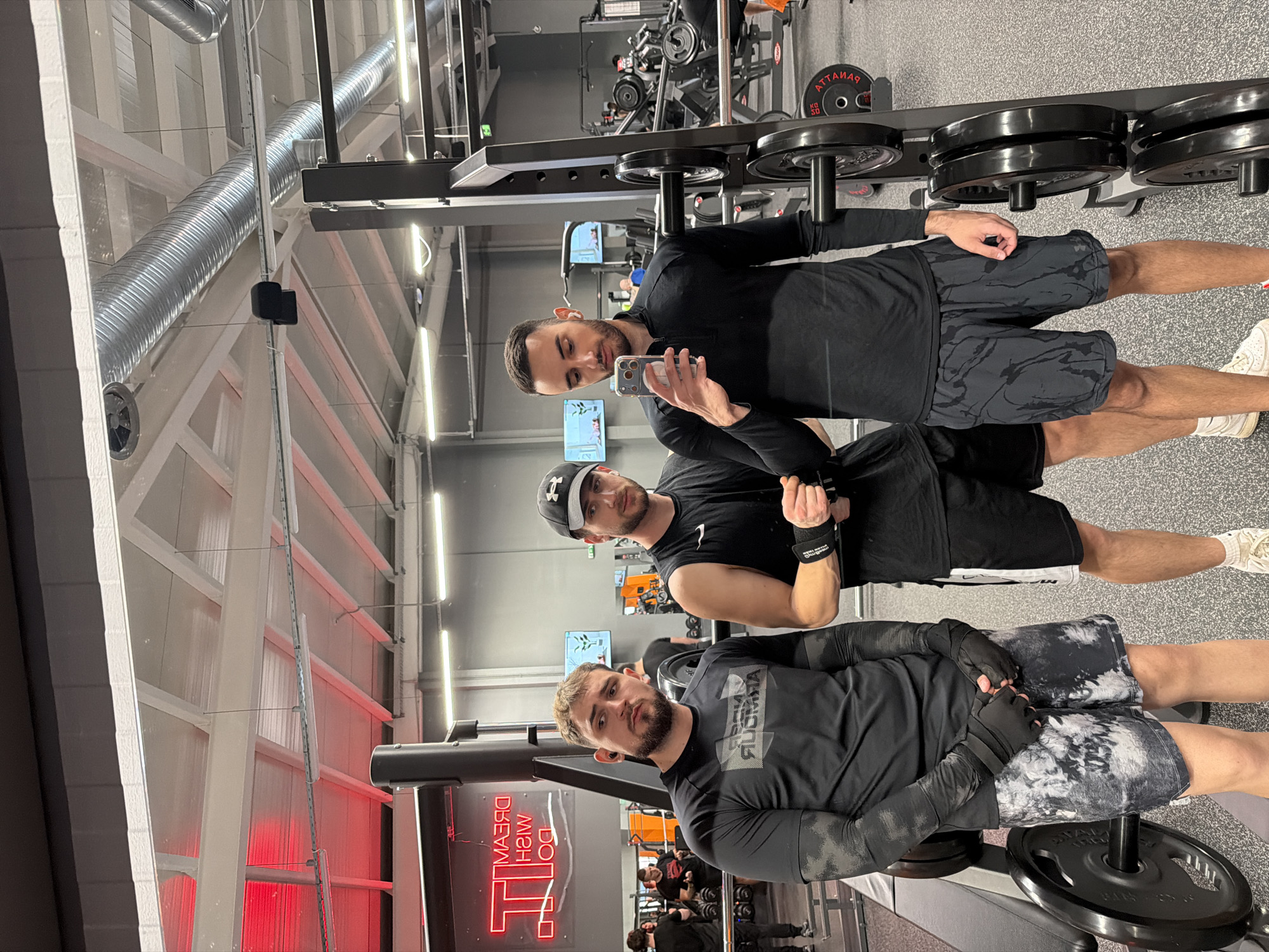 Three men are posing for a selfie in a gym. The gym has weightlifting equipment, including weight racks and dumbbells. In the background, other people are working out. The gym has white walls, white beams, and metal ventilation ducts. A sign that reads "Dream, Wish, Do It" in red neon lights is visible on the wall. It is not possible to determine if the location is in Plovdiv, Bulgaria from the image.
