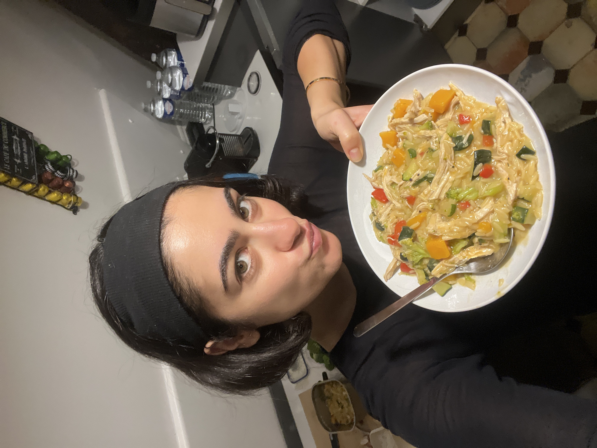 A woman is holding a white plate filled with cooked pasta, shredded chicken, and various vegetables, including zucchini, red bell peppers, and butternut squash. She is wearing a black long-sleeved shirt and a black headband. A gold bangle is on her wrist. The background shows a kitchen setting with a countertop, a spice rack, and bottled water. The floor has a patterned tile design. Although the photo shows elements suggestive of Paris (French signage, tile style) more definitive evidence is required to determine the location with certainty.