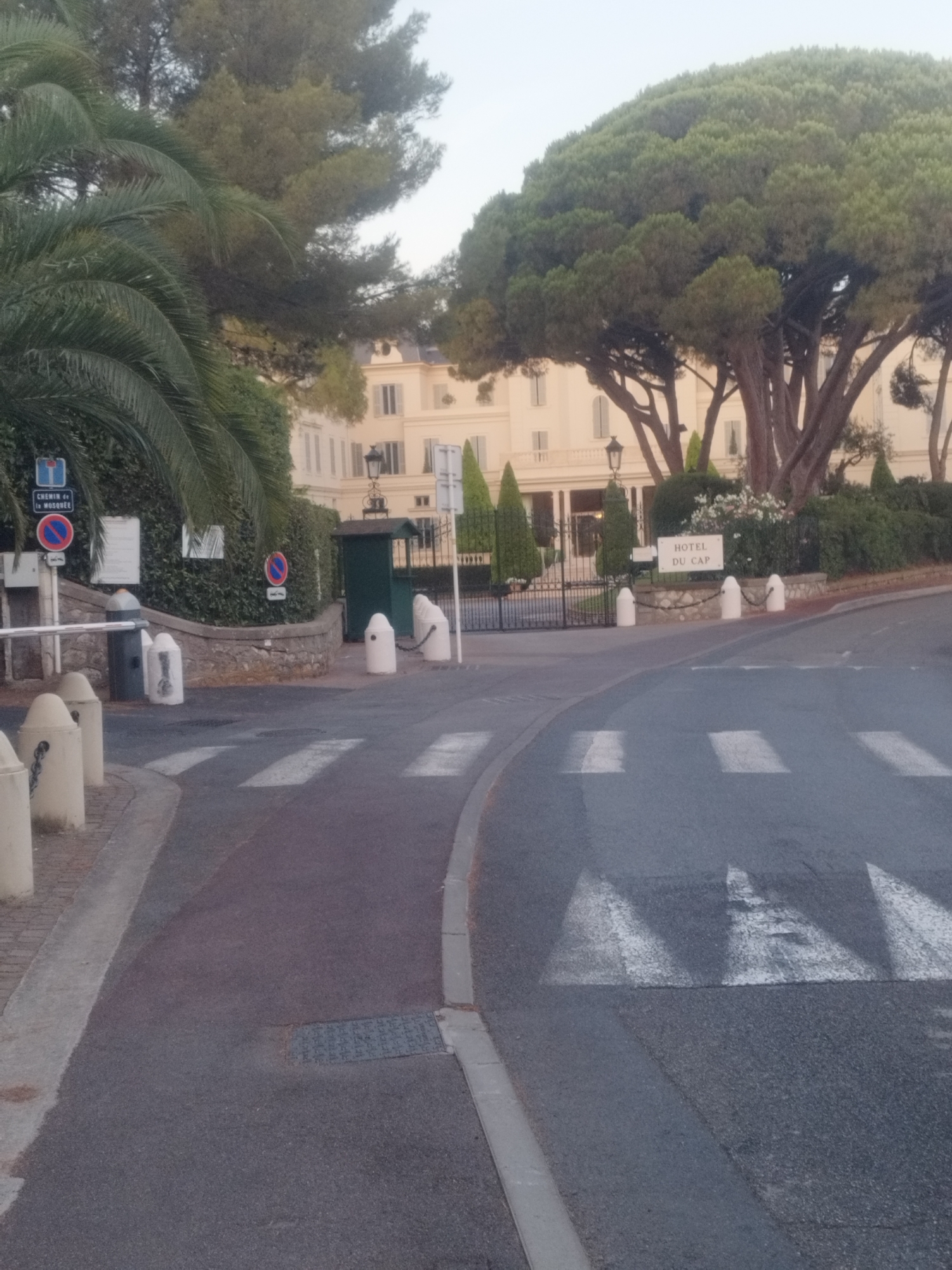 The image captures an entrance to the Hotel du Cap-Eden-Roc in Antibes, France. A paved road with a pedestrian crossing is in the foreground, leading towards a grand entrance. The hotel itself is a large, light-colored building with numerous windows and a classic architectural style, partially obscured by lush green trees, including a large pine tree with a distinctive shape and a palm tree on the left. A gate marks the entrance to the hotel grounds, and a white sign reads "HOTEL DU CAP". There are bollards and chains along the roadside and at the entrance, suggesting a controlled access area. A small guard booth is visible near the gate. The overall atmosphere is one of elegance and exclusivity, set within a well-manicured natural environment.