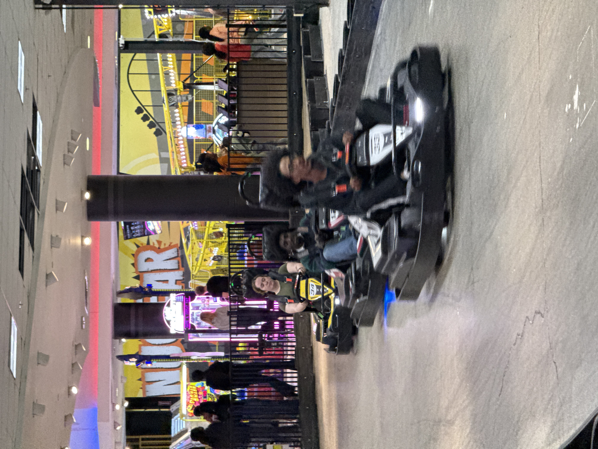The image depicts a go-kart track at an entertainment venue, likely in Columbus, United States of America. Several go-karts are racing, each occupied by a driver. The karts are in motion, indicated by blurring. The background includes arcade games and other entertainment attractions, with various colorful lights and signage. A metal barrier separates the track from the arcade area. Pedestrians are observing the race from behind the barrier. The flooring of the track is a grey surface.