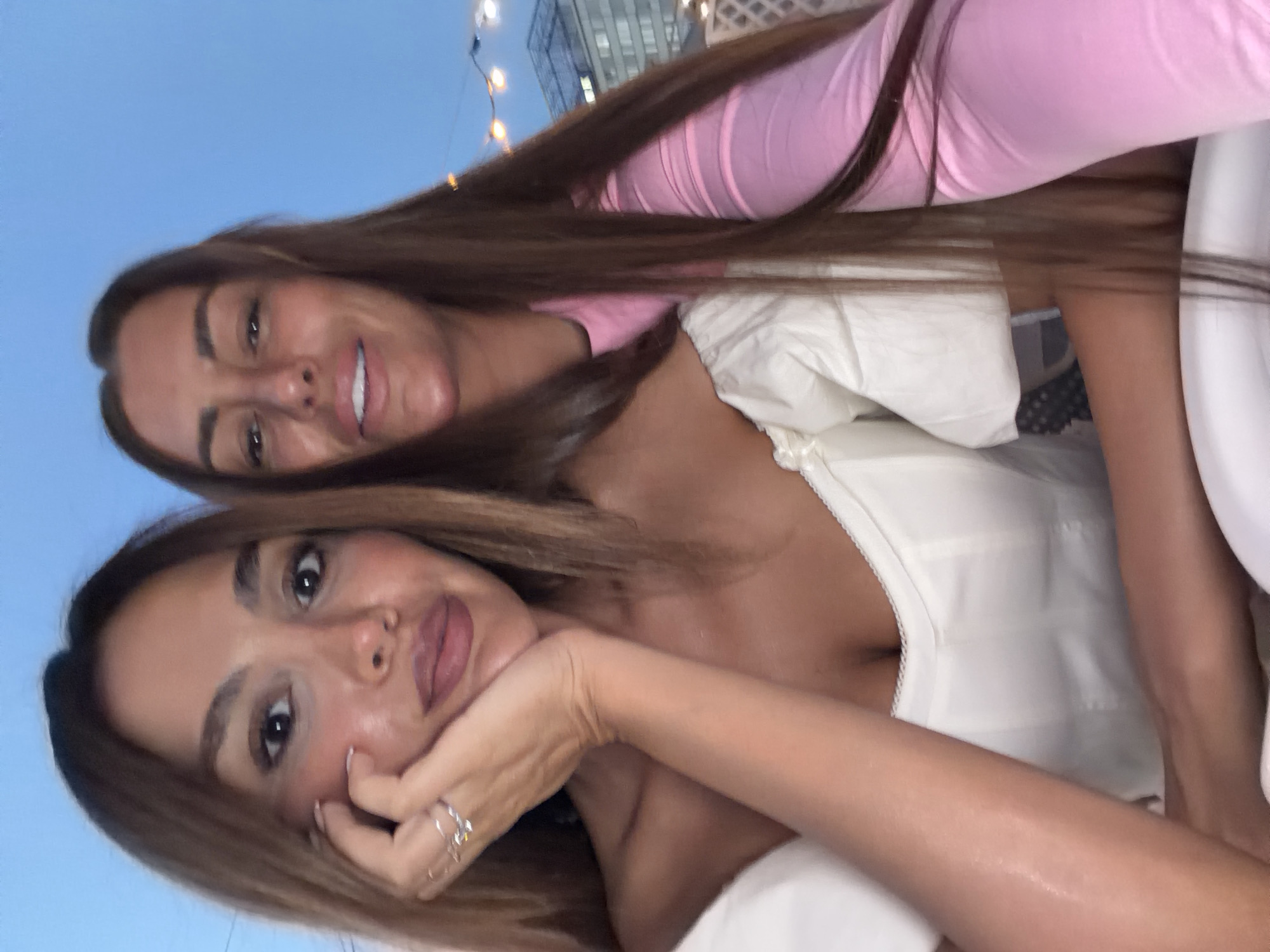 Two women are captured in a selfie, with the woman on the left in the foreground and the woman on the right in the background. Both have long, brown hair. The woman on the left is wearing a white, off-the-shoulder top. Her left hand, adorned with rings, is resting on her chin. The woman on the right is wearing a pink long-sleeved top. They appear to be outdoors, with a dusky blue sky visible above. In the background, out-of-focus string lights and a building are visible, suggesting an urban setting. The image does not contain any information that would indicate the location is in Novosibirsk, Russia.