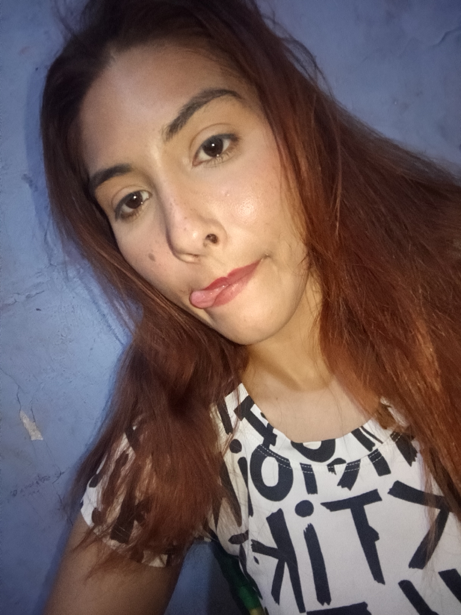 A young woman poses for a selfie. She is wearing a white shirt with black letter-like shapes printed on it. Her long auburn hair frames her face. She sticks her tongue out slightly. She has light skin and brown eyes. The background appears to be a textured blue wall. The location, Juan Griego, Venezuela, is not verifiable from the image content.