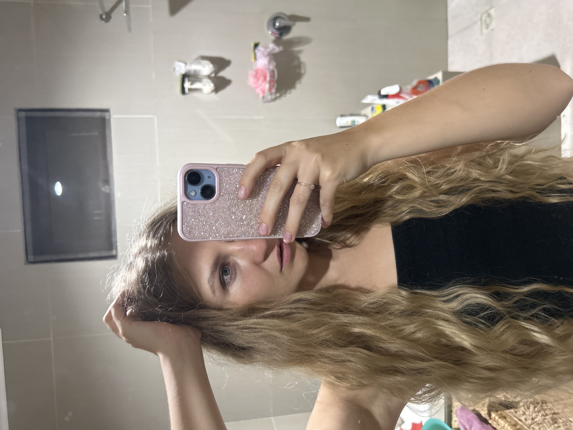 A mirror selfie captures a young woman in what appears to be a bathroom in Kuta Selatan, Indonesia. The image is presented vertically but suggests a sideways capture, with the woman holding her phone in her left hand to take the photo.
The central subject is a woman with long, wavy blonde or light brown hair that cascades down her shoulders. Her face is partially visible, showing one blue eye and her lips with a neutral expression. She is wearing a dark top, possibly black. Her left hand, adorned with a delicate ring featuring a small stone on her ring finger, holds an iPhone encased in a sparkly pink protective cover. Her right hand is seen reaching towards her head, touching her hair.
The background, reflected in the mirror, reveals typical bathroom elements. These include light-colored tiled walls and fixtures such as a shower head. Various toiletries are visible, including a pink loofah or bath sponge and several bottles (likely shampoo, conditioner, or lotions) placed on a shelf or ledge. A rectangular frame, possibly part of a shower enclosure or another mirror, is faintly visible on the left side of the reflection. The lighting suggests an indoor setting, likely with artificial illumination, so the time of day is not evident. There is no discernible text visible within the image.