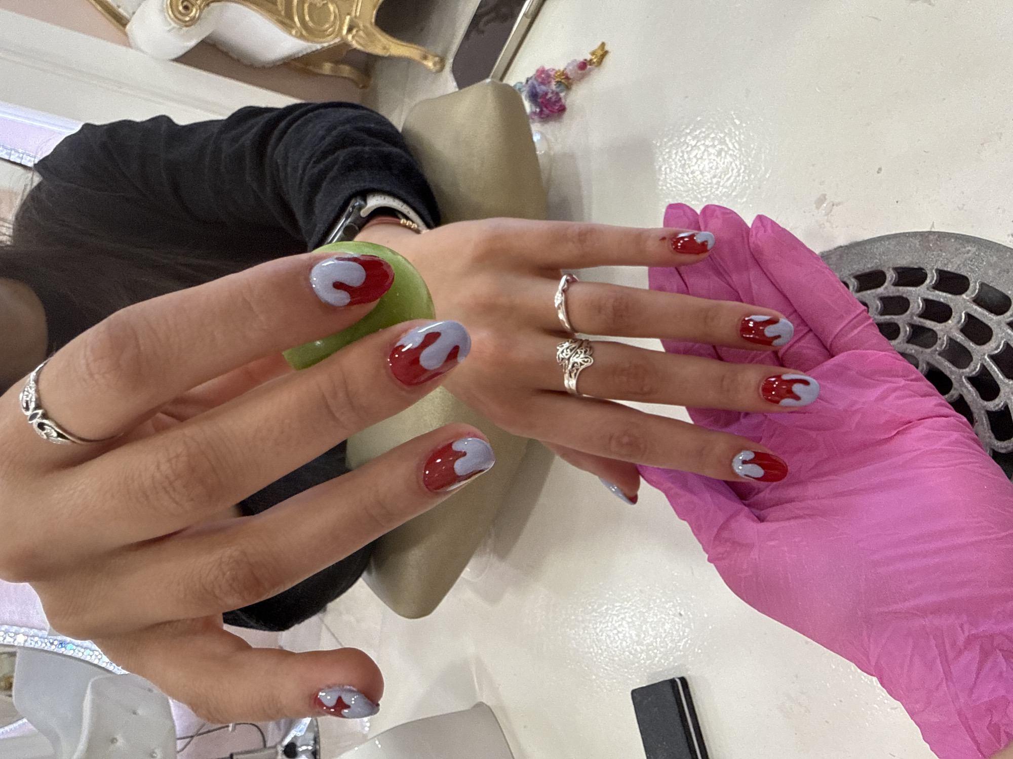 The image shows a woman's hands with a fresh manicure. The nails are short and painted red with a light purple accent. The woman is wearing three rings on her fingers: one on the middle finger, and two on the ring finger. She is holding a green apple in one hand. One hand is resting on a pink gloved hand, presumably a nail technician. The background shows a manicure table with various tools and equipment. It is impossible to determine the location in Sofia, Bulgaria from the image.
