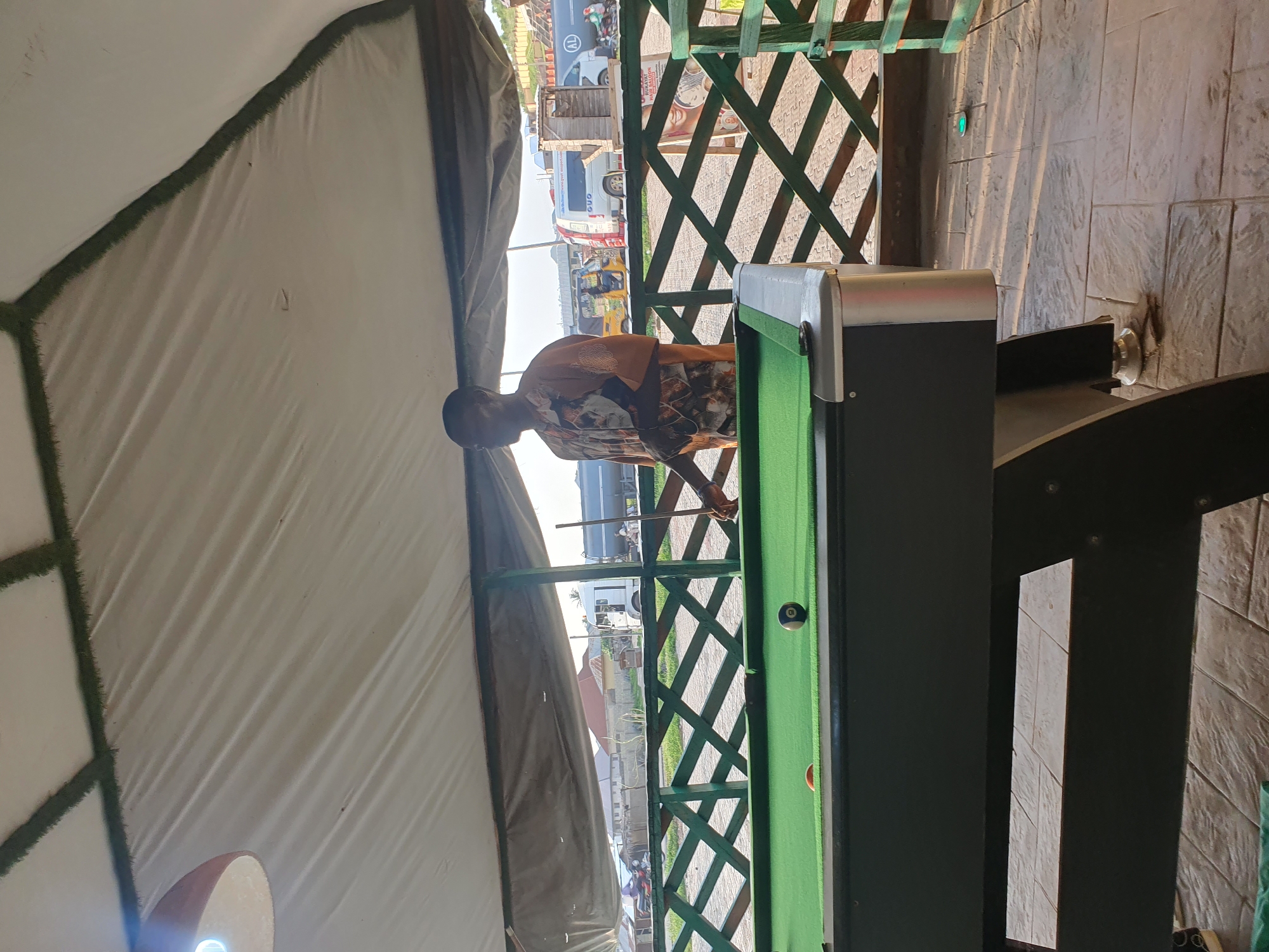 In Lokoja, Nigeria, a man stands on a paved patio next to a green billiards table. He is wearing a short-sleeved, patterned shirt and appears to be preparing to play. The billiards table has several balls on it, including an 8-ball. Behind the table, a green wooden fence with a diamond pattern separates the patio from a road. Several vehicles, including trucks and a motorcycle, are visible on the road. The scene is shaded by a large, white tent. The time appears to be late afternoon or early evening, based on the angle of the sunlight.