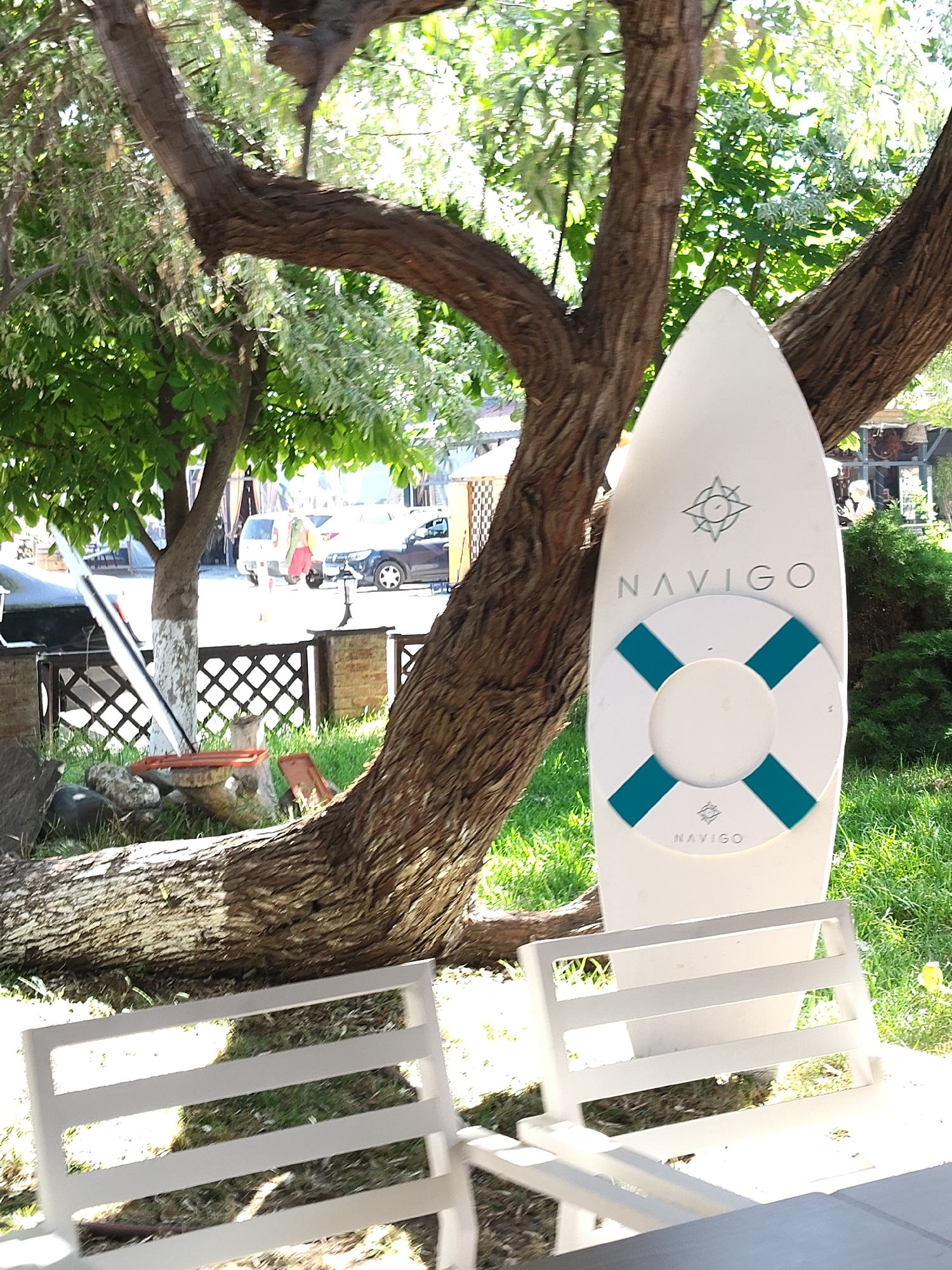 An outdoor scene depicts a surfboard-like object leaning against a tree trunk, featuring the "NAVIGO" logo and a life preserver graphic. The surfboard is white with teal accents on the life preserver. In the background, a fence made of dark wood lattice separates the foreground from a paved area with parked vehicles, including a white van and a dark car. Two people are visible near the van, one wearing red pants and carrying a light-colored garment. The environment consists of green grass, trees with textured bark, and dappled sunlight filtering through the leaves. The scene suggests a recreational or hospitality setting.