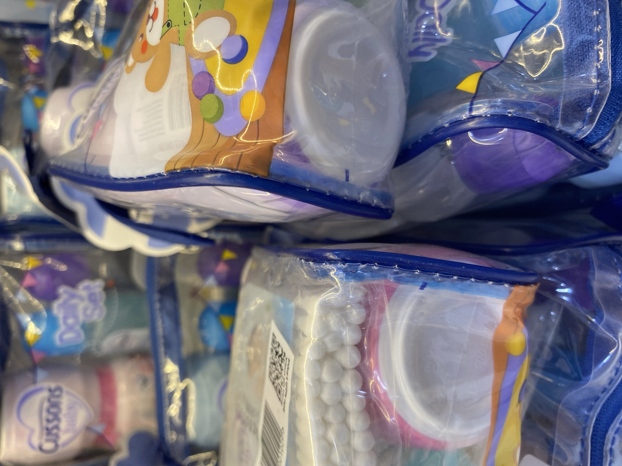 The image displays a close-up, eye-level view of several retail product packages, likely baby care items, stacked and partially obscuring one another. The packaging is predominantly transparent plastic, revealing the contents within.
On the left side, a package features pink and white branding with the text "Cussons Baby". Below this, another package shows rows of white cotton swabs. Further down, a pink and white circular item is visible.
The center and right side of the image are dominated by packages with colorful graphics. One package prominently displays a cartoon bear wearing a green outfit, along with various colored circles and abstract shapes. Another package visible towards the bottom right has a blue zipper and a light blue graphic with some text that is partially legible as "Winni".
The overall impression is one of a retail display, possibly in a supermarket or specialty store. The lighting appears to be artificial, creating reflections on the plastic packaging. There are no people or animals visible in the image. The background is blurred, suggesting the focus is entirely on the product packaging.
There is no information in the image to suggest a specific location in Lamteumen Barat, Indonesia.