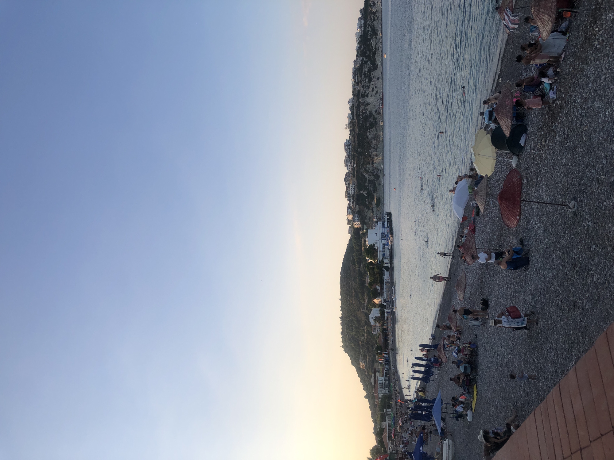 The image depicts a crowded pebble beach in Karaburun, Turkey, at dusk. The sky is a clear, pale blue, transitioning to a pale yellow near the horizon. The sea is calm and reflects the soft light.
Numerous people are visible on the beach, engaged in various activities. Some are sitting under umbrellas, others are walking along the shore, and a few are in the water. Beach umbrellas, predominantly in shades of blue, yellow, and red, dot the landscape, providing shade for the beachgoers. Beach chairs and towels are scattered across the pebbles.
In the background, a hilly landscape rises from the coast, dotted with white buildings and greenery. A few boats are anchored in the bay, and a larger structure, possibly a pier or a waterfront building, is visible on the left side of the image. The overall scene conveys a lively and relaxed atmosphere typical of a summer evening at a popular beach destination.