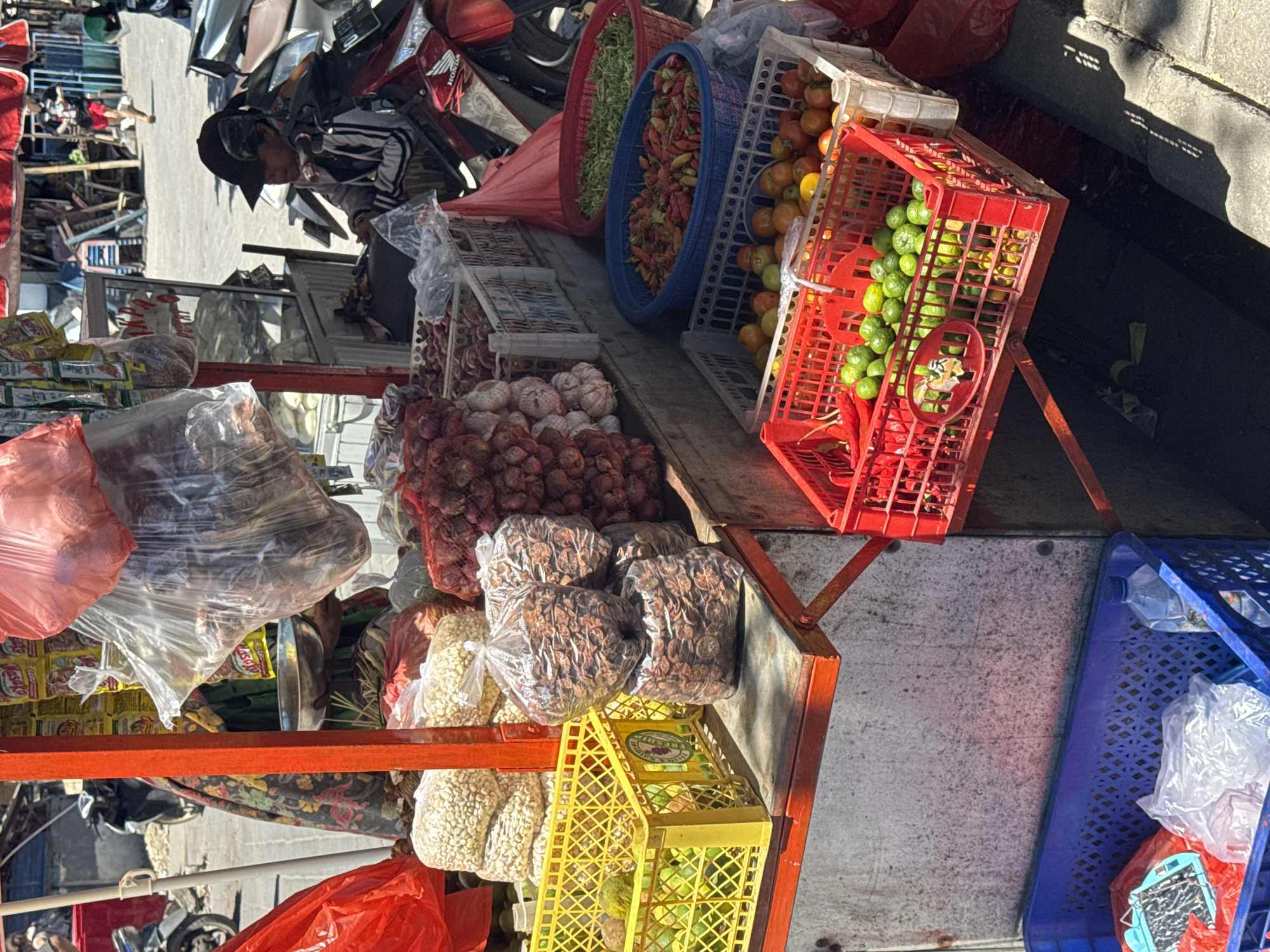 The image shows a street market scene in the city of Unknown, Unknown. A vendor is partially visible, wearing a hat and patterned shirt. A motorcycle is parked nearby.
The market stall displays a variety of produce, including onions, garlic, nuts in plastic bags, green limes and oranges in red and white plastic crates, green beans, and chili peppers in blue plastic tubs. Yellow plastic crates hold additional items. The produce is arranged on a metal countertop. Other plastic bags, some red, some clear, also occupy the scene. The ground is concrete.