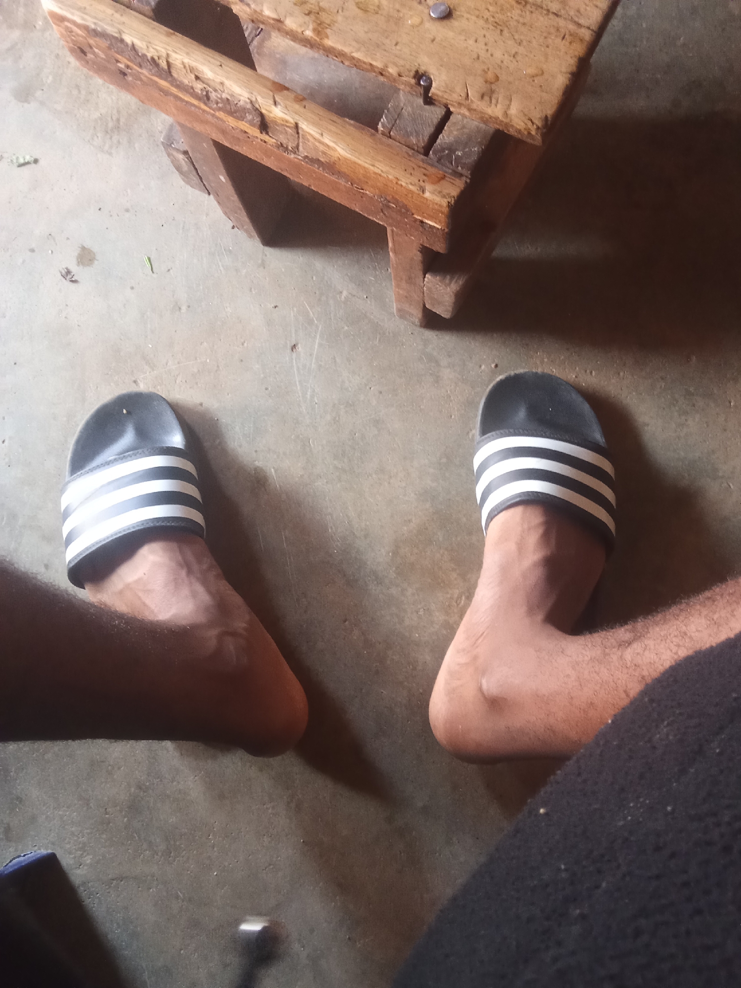 The image shows a close-up view of two feet wearing black and white Adidas slide sandals. The feet are on a concrete floor. In the background, a rustic wooden stool or bench is visible. The lighting suggests an indoor or shaded outdoor environment. The scene appears to be a casual, everyday setting. No specific location in Jos, Nigeria can be determined from the image.