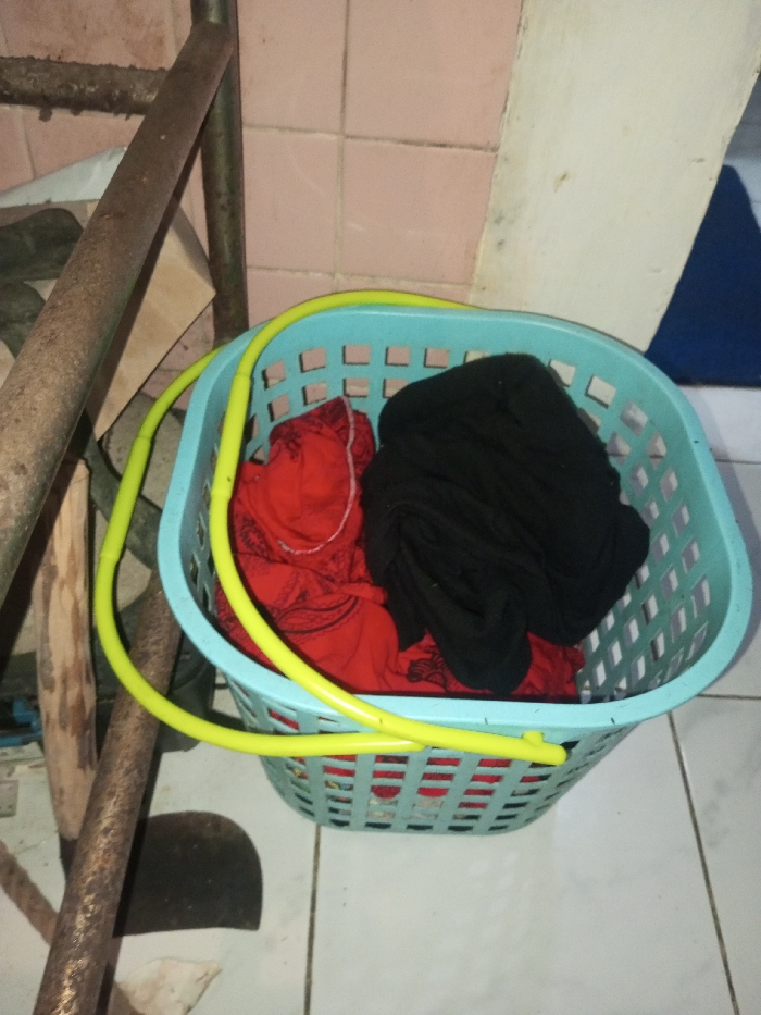 A light blue laundry basket with a yellow ring around its opening sits on white tiled floor. Inside the basket are crumpled piles of red and black clothes. To the left, a rusty metal structure stands beside the basket. The background features pink tiled walls, white walls, and a glimpse of a blue object. The scene suggests a laundry area or utility room.