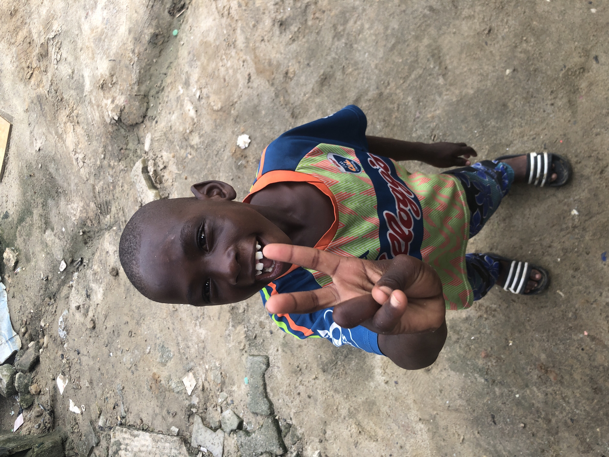 In a dirt-covered outdoor area in Ado, Nigeria, a young Black boy stands looking directly at the camera. He is wearing a blue, green, and orange t-shirt with a white graphic and pattern, and blue shorts with a floral pattern. On his feet are black sandals with white stripes. The boy is holding up his right hand, with his index and middle fingers extended in a peace sign. His mouth is open, revealing missing front teeth, and he appears to be smiling. The ground around him is rough and uneven, with scattered debris and patches of mud. The overall impression is one of a candid, everyday moment captured in a somewhat unkempt urban environment.