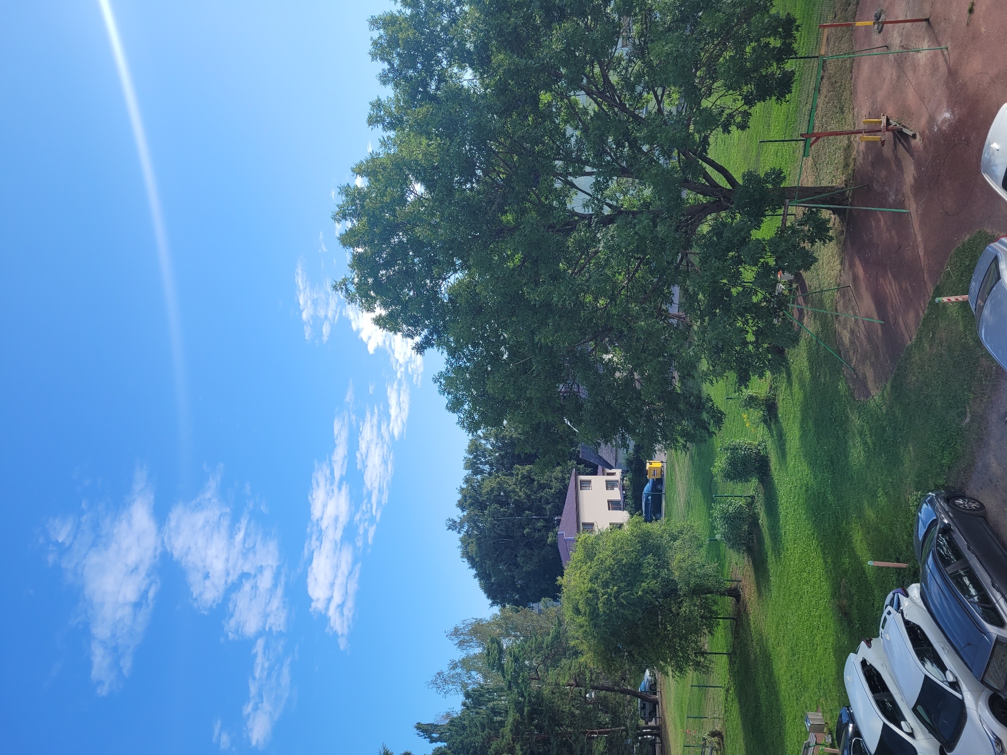 The image captures a serene outdoor scene, likely in a residential area, with a bright blue sky dotted with wispy white clouds and a strong sunbeam extending across the upper right portion. In the foreground, a paved area shows several parked cars, including a dark grey sedan and two white SUVs. A narrow dirt path with a red-and-white striped bollard is also visible.
Beyond the parked cars, a lush green lawn stretches out, bordered by a metal fence. Scattered across the lawn are several trees of varying sizes and foliage density. A prominent, large deciduous tree with a full canopy dominates the mid-right of the frame, its branches casting shadows on the ground. At the base of this large tree, there is a rudimentary playground structure with two swing sets.
Further back, a light-yellow, two-story building with a brown roof is visible amongst a cluster of trees. Another building with a dark roof is partially visible to the right of the yellow building. In the background, a dense line of evergreen and deciduous trees forms a natural boundary. The overall impression is one of a peaceful, well-maintained suburban or community outdoor space on a sunny day, consistent with a location in Jūrmala, Latvia, known for its residential areas and natural landscapes. No people are visible in the image.