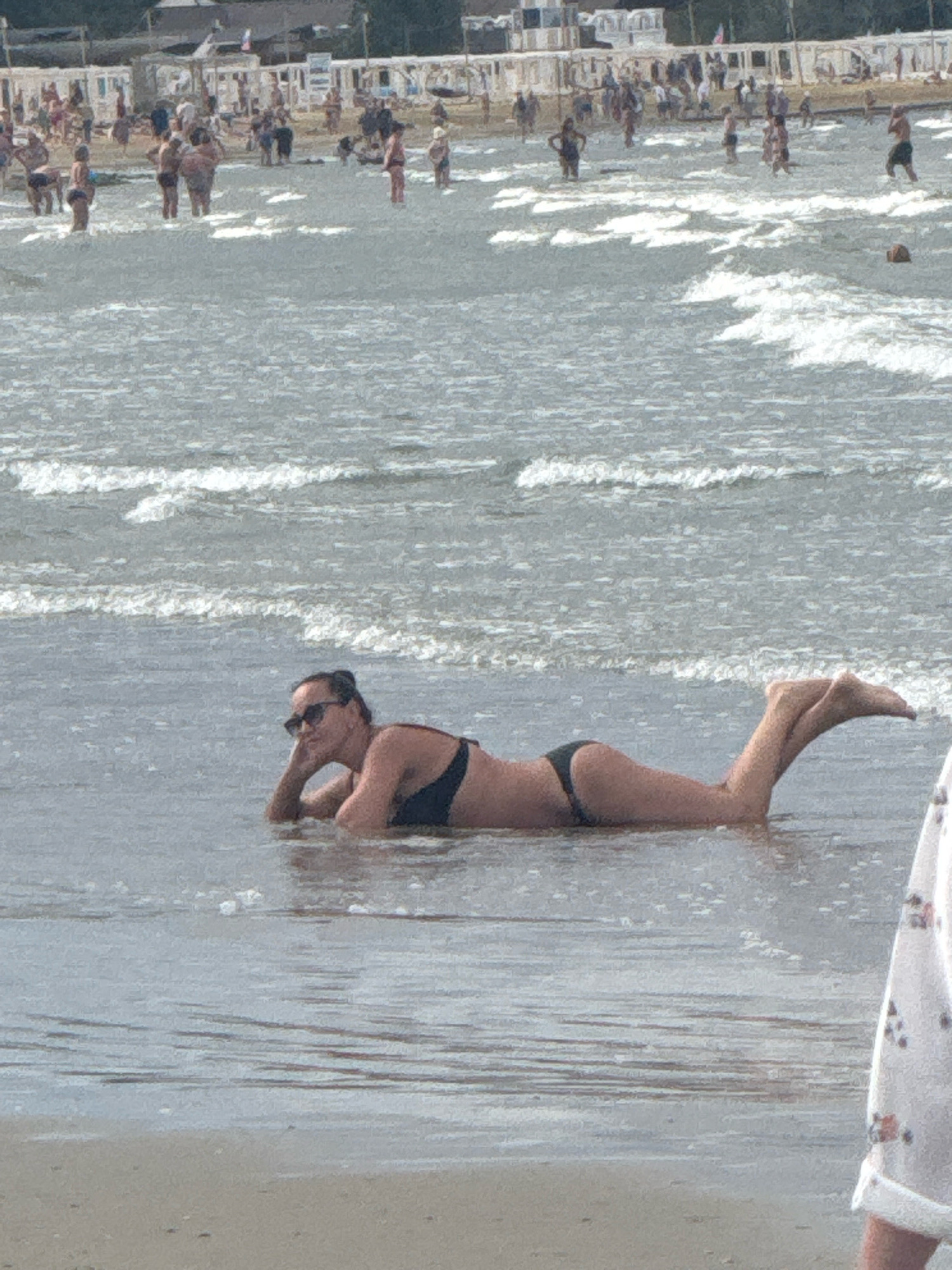A woman in a black bikini is lying on the wet sand at the edge of the sea, facing left. Her head is propped up on her left hand, and her right arm is bent behind her. She is wearing sunglasses. The water is shallow around her, with small waves breaking. In the background, other beachgoers are visible, some standing in the water and others on the sandy shore. Behind them, there are rows of white beach huts or cabanas. The scene depicts a typical day at a beach resort, possibly on the Black Sea coast of Anapa, Russia, given the architectural style of the beach huts.