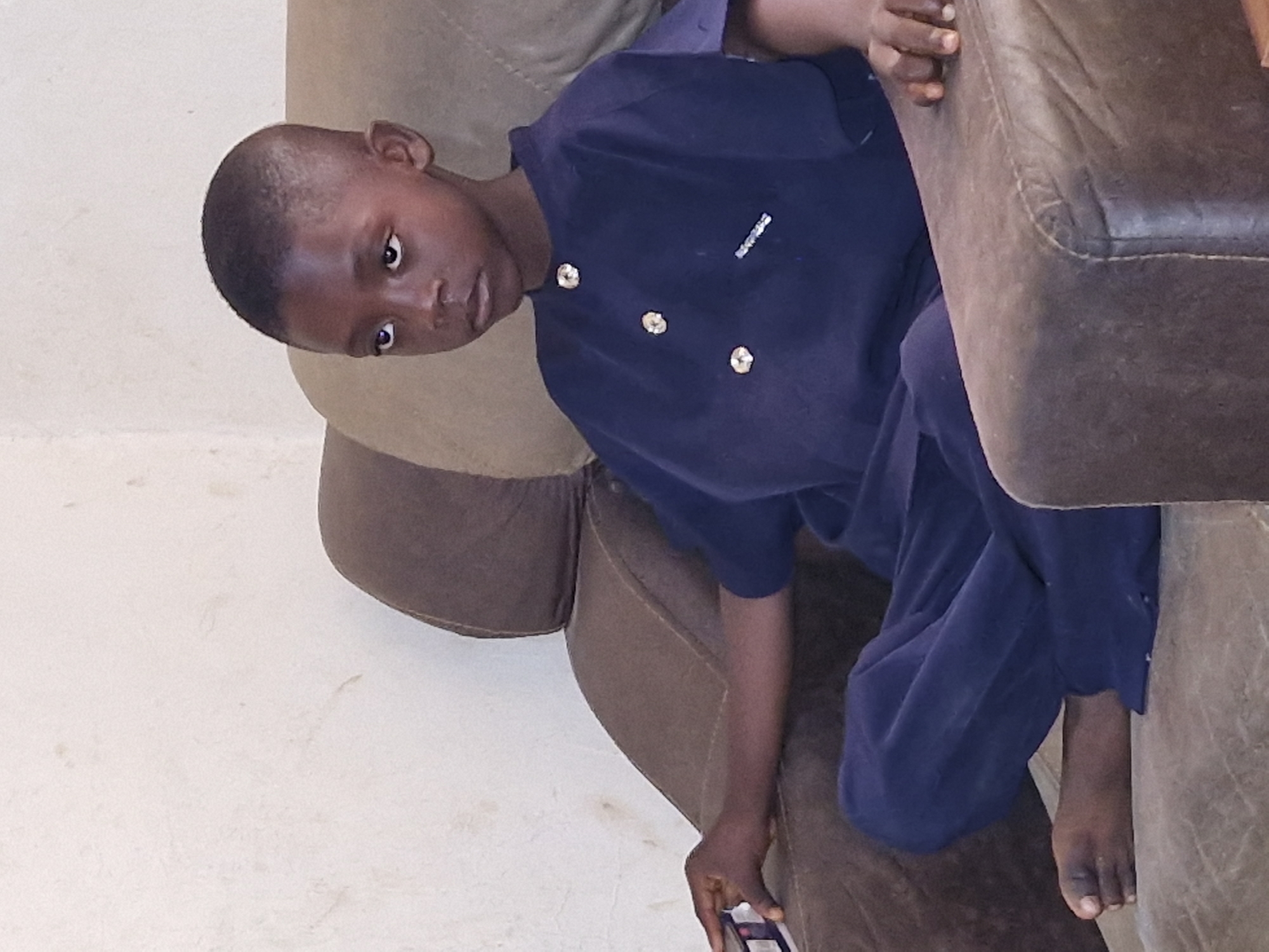 A young Black boy sits on a plush, brown armchair, facing slightly to the left. He is wearing a dark blue, collared tunic with three large, iridescent buttons and matching dark blue pants. His left arm rests on the armrest of the chair, and his left hand holds a small, rectangular object with a white and purple label, possibly a box of medication. His right hand is not fully visible. His bare feet are visible, with his left foot resting on the seat of the chair. The boy's eyes are wide and directed upwards and to the left, suggesting he is looking at something out of frame. The background is a plain, textured light-colored wall. The lighting is soft, casting subtle shadows. The overall impression is of a child engrossed in something unseen.
