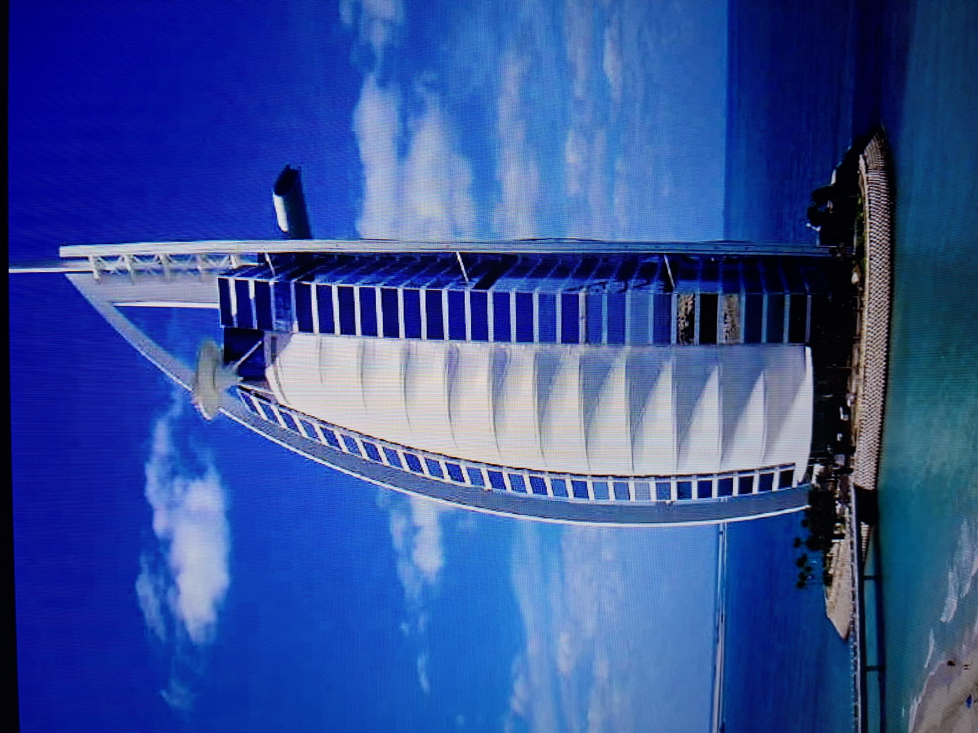 The image shows the Burj Al Arab hotel in Dubai. The hotel is situated on an artificial island near the shore of the Persian Gulf. It is distinguished by its sail-like structure, white curved facade, and a tall mast-like element at the top. The surrounding environment includes the blue sky with scattered clouds and the turquoise water of the Gulf. There is a land connection visible along the shore of the man made island of the hotel.