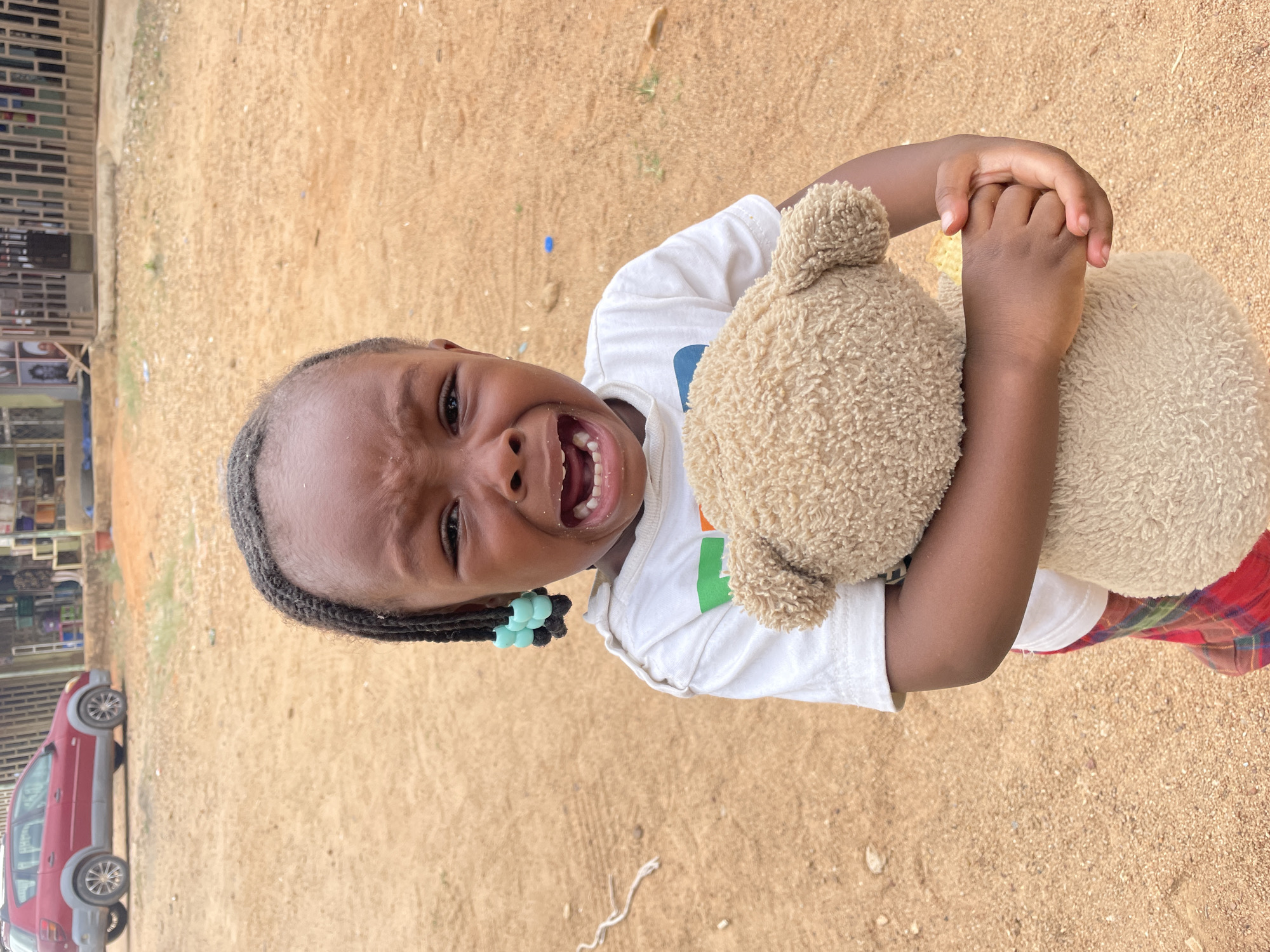 A young child, likely between 2 and 4 years old, is crying and holding a stuffed teddy bear. The child has dark skin, braided hair with blue beads, and is wearing a white t-shirt and a plaid garment. The background consists of sandy ground. A building with some open spaces and a car are visible in the distance. This image appears to have been taken outside in Barakin Akawo, Nigeria.
