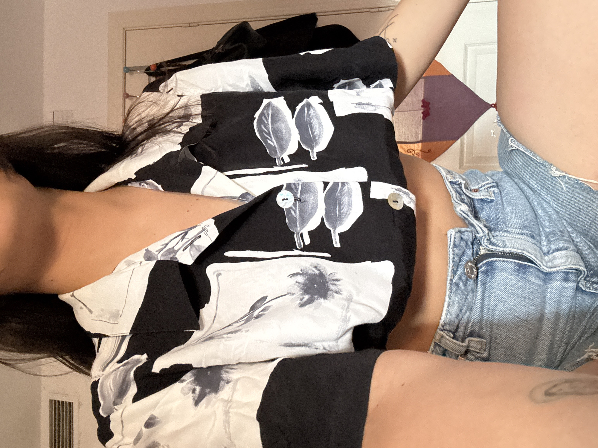 The image shows a close-up, medium shot of a person wearing a black and white floral print crop top and light wash denim shorts. The person has long dark hair and is sitting down. The background includes a white door and a decorative kite. The scene is indoors and does not provide any information about the city of Las Rozas de Madrid, Spain.