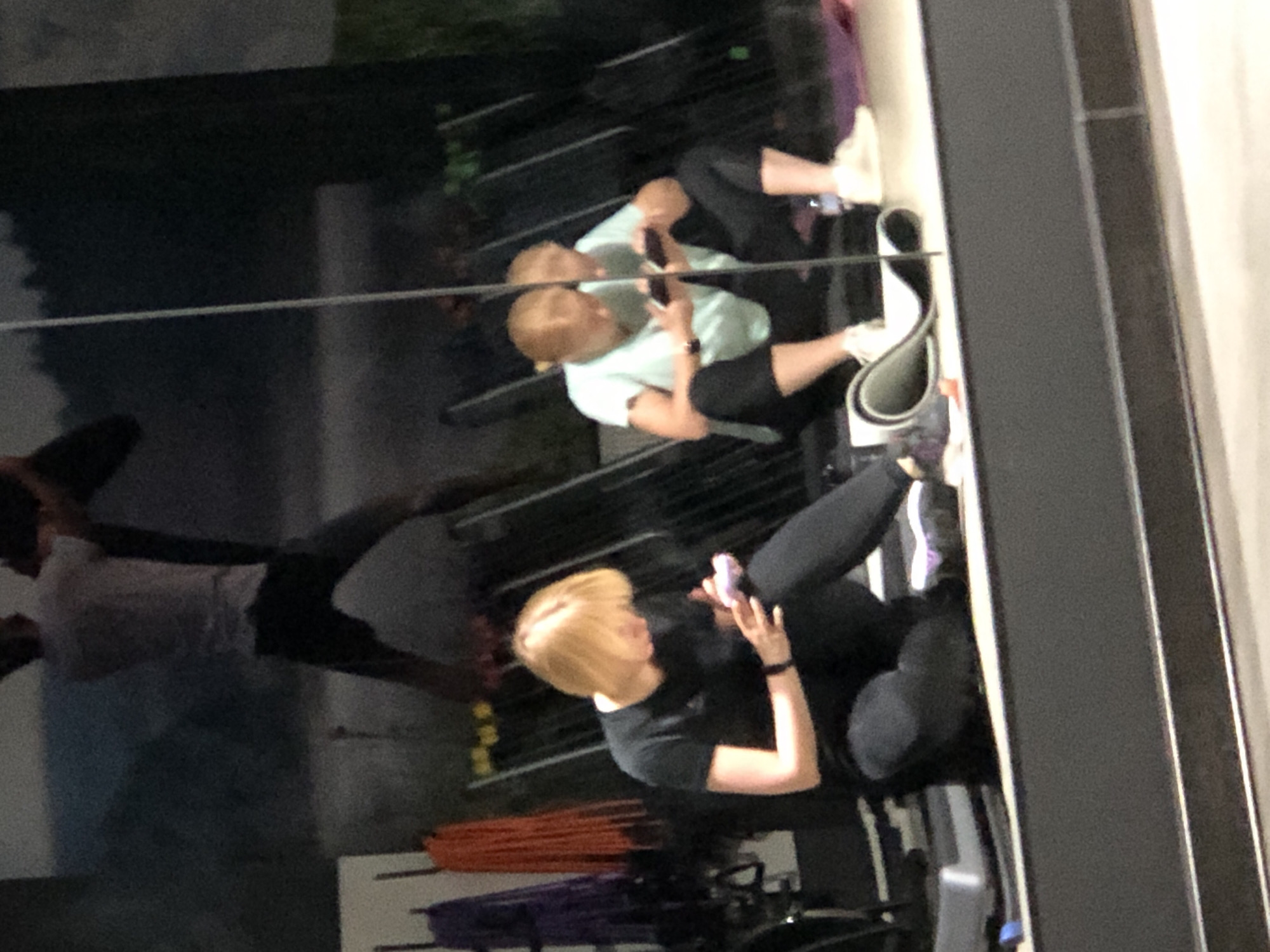 The image shows two young women in a gym, possibly a fitness studio, near the city of Saint Petersburg, Russia. One woman, with blonde hair, is wearing a black t-shirt and black leggings. She is sitting on the floor, looking down at her phone, which she is holding in her right hand. She is also holding a pink object in her left hand. The other woman, with reddish-blonde hair, is wearing a light blue t-shirt and black leggings. She is also sitting on the floor, in a squatting position, looking at her phone. Her reflection is visible in a large mirror behind her, which also shows a blurry image of a man in the background, appearing to be exercising. The gym appears to have workout equipment, including racks of resistance bands or ropes in shades of purple and orange hanging on the wall. A yoga mat is partially visible on the floor. The lighting suggests it might be evening or indoors.