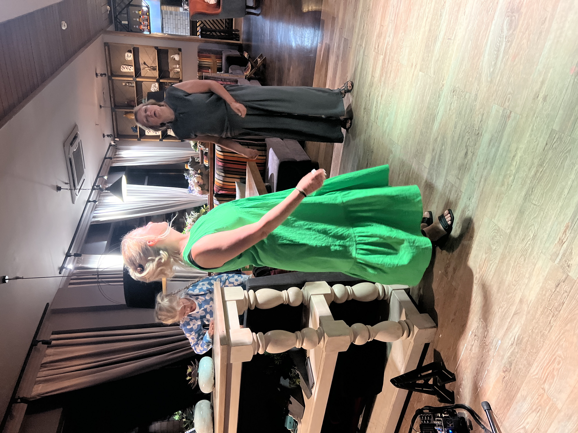 An indoor scene in what appears to be a restaurant or event space features three people and various objects. In the foreground, a young girl with blonde hair in a ponytail, wearing a blue and white patterned long-sleeved shirt, leans over a wooden table with decorative legs. To her right, a woman with blonde hair, facing away from the camera, is wearing a bright green, sleeveless, tiered dress. Further to the right, a woman with short blonde hair, wearing a dark sleeveless top and wide-leg pants, stands facing the other two individuals, looking upwards as if speaking. Behind the table, a dark curtain hangs, and to the right, shelves filled with decorative items and bottles are visible, along with seating arrangements. The floor is made of light-colored wood planks. On the left side of the image, a piece of audio equipment is placed on the floor near a black stand. The lighting suggests an evening or dimly lit indoor setting, with some colored light effects on the floor. The scene is located in Novosibirsk, Russia.