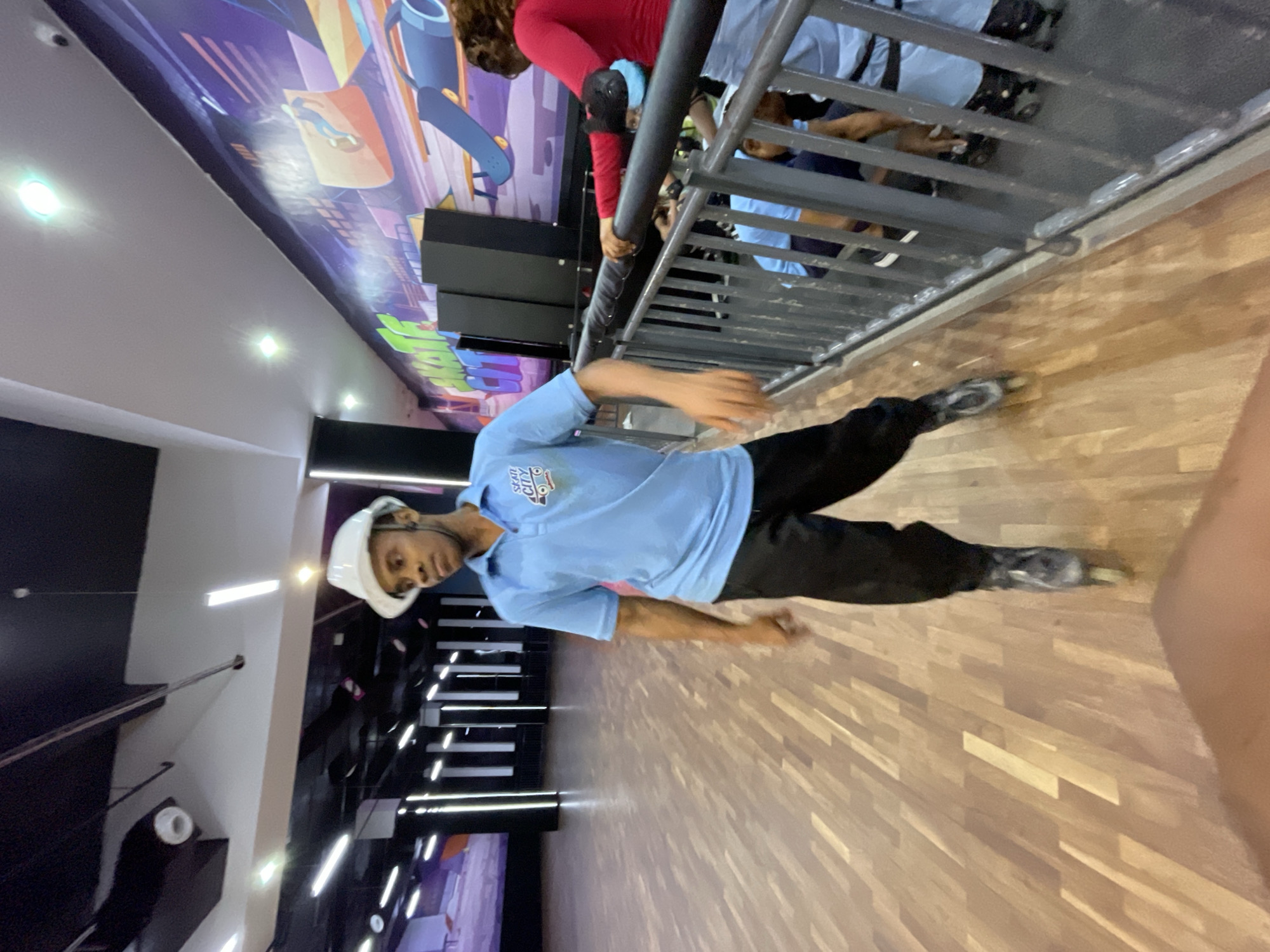 A young Black male wearing a white protective helmet, a light blue polo shirt with a "SKATE CITY" logo, and black pants with inline skates on his feet is the central subject in the foreground of this indoor skating rink. He is facing to the right of the frame, with his left arm bent and his right arm extended slightly forward, suggesting movement. The skating rink features a polished wooden floor and is illuminated by strip lights. In the background, a large, colorful mural depicting skateboards and abstract designs adorns the wall. To the right, a metal railing separates the skating area from a seated section where other individuals are present, including a Black female in a red long-sleeved shirt and elbow pads, and several other people wearing blue shirts. The environment appears to be a recreational facility, likely a roller skating or rollerblading rink. The overall impression is that of an active and vibrant public space for skating.