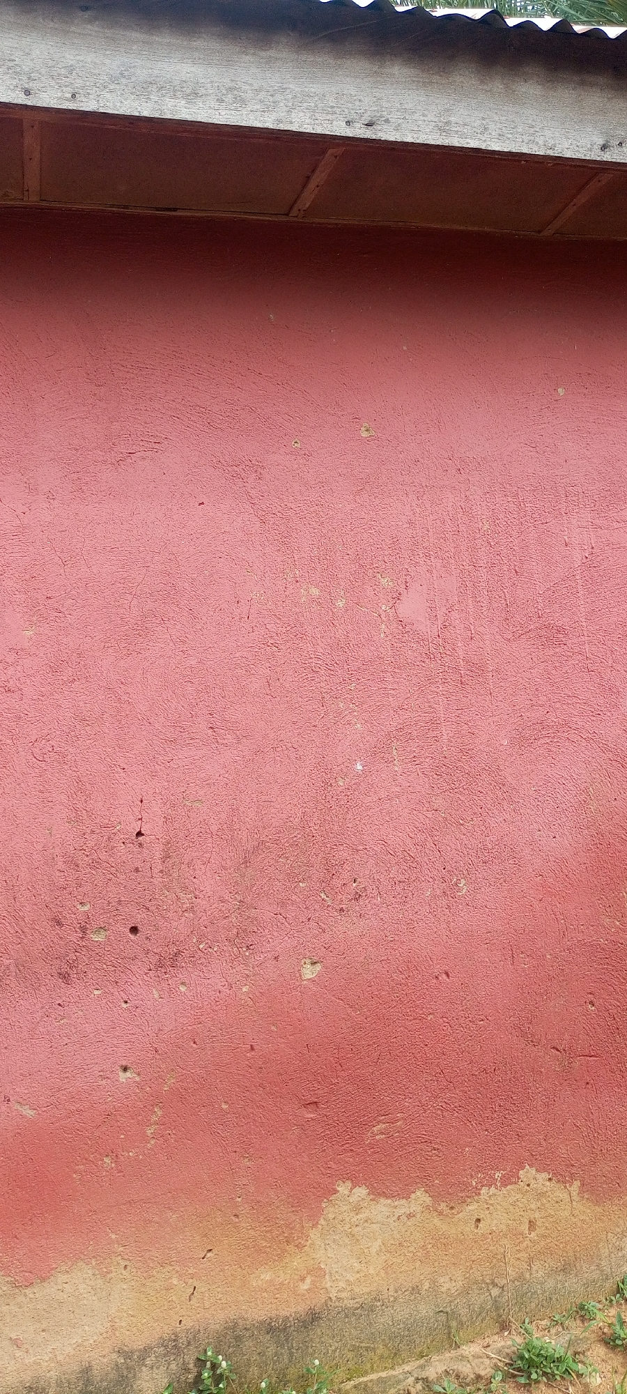 The image shows the side of a building with a red, textured wall. The wall has several imperfections, including cracks and small holes. At the top of the image, there is a wooden eave and a corrugated metal roof. The bottom of the wall transitions into a lighter, dirtier color, suggesting it is closer to the ground. Some sparse green grass is visible at the very bottom. The scene appears to be an outdoor setting, likely in a residential or commercial area. The style of construction suggests it could be located in Jalingo, Nigeria.