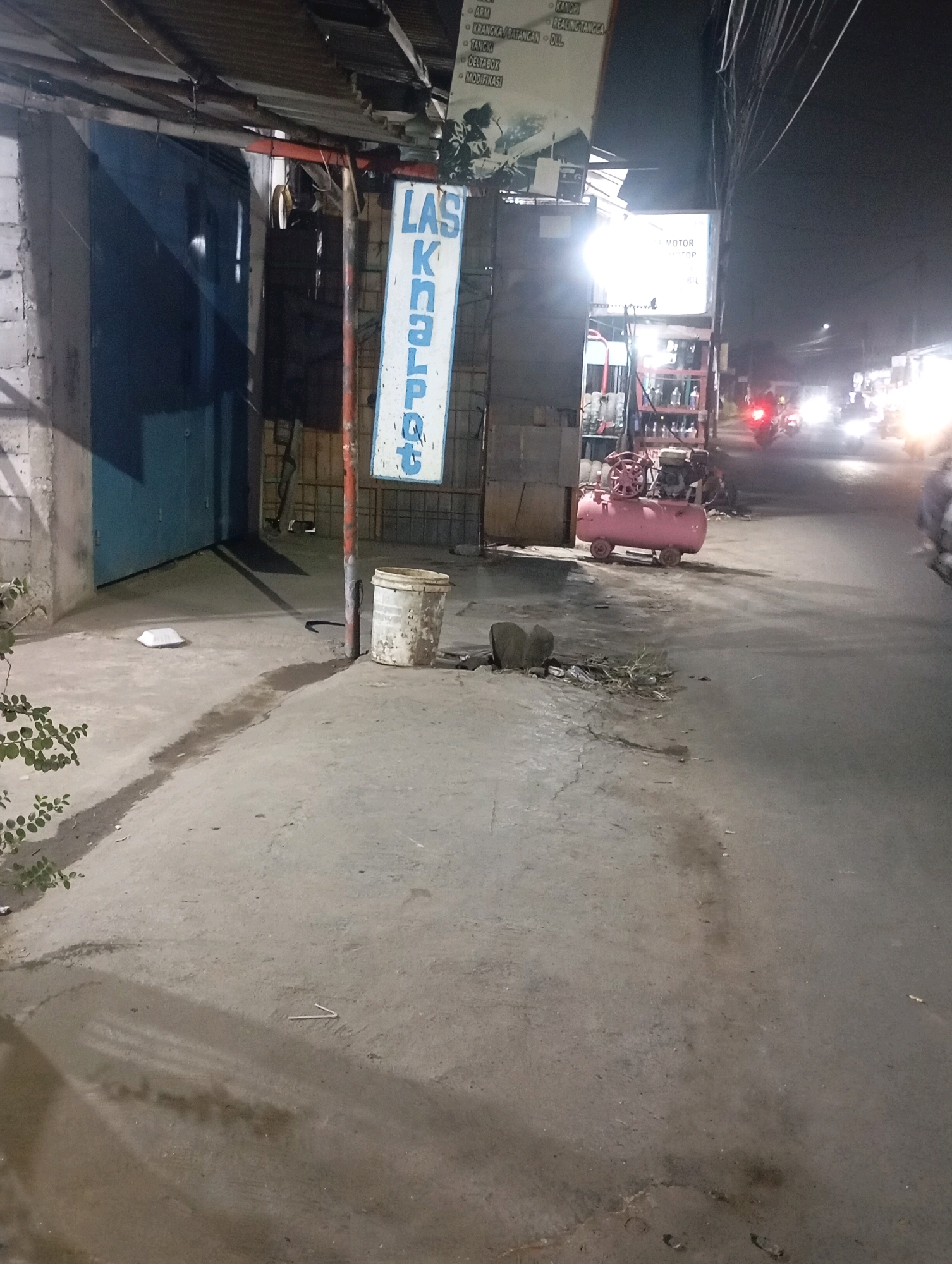 The image depicts a nighttime street scene in Babelan, Indonesia. A storefront with a blue garage door is visible on the left, with a sign above it that reads "LAS KNALPOT" in blue lettering on a white background. To the right of the storefront, a pink air compressor sits on a dirt and asphalt surface. Further down the street, traffic lights and headlights of vehicles illuminate the dark road. In the foreground, a white plastic bucket is placed on the ground next to a metal pole. The overall environment suggests a commercial area, possibly a repair shop, on a typical night in Babelan.